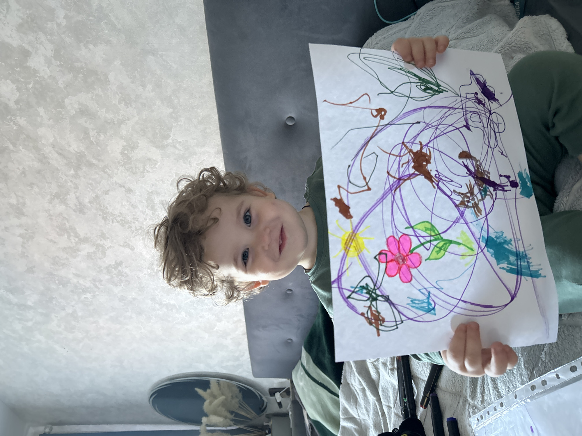 A young child with light skin, blue eyes, and curly light brown hair is positioned centrally, seated on a bed in what appears to be a bedroom in Klin, Russia. The child, wearing a green long-sleeved shirt and green pants, holds up a white sheet of paper displaying a colorful drawing. The drawing features a prominent pink flower with a green stem and leaf, a yellow sun with rays, and abstract scribbles in purple, brown, blue, and green. The child exhibits a smiling expression, looking directly towards the viewer.
In the background, a light-colored, textured wall is visible. To the left, a portion of a dark-rimmed oval mirror reflects light-colored dried decorative plants in a vase, and a blue window frame with a curtain is partially visible. The bed features a grey upholstered headboard with a single visible tuft and light-colored, textured bedding. A green pillow rests on the bed to the child's left. On the bed surface in the lower left, several dark-colored markers are scattered alongside a clear plastic sheet protector. A fluffy, light-colored blanket or garment is visible on the bed to the child's right.