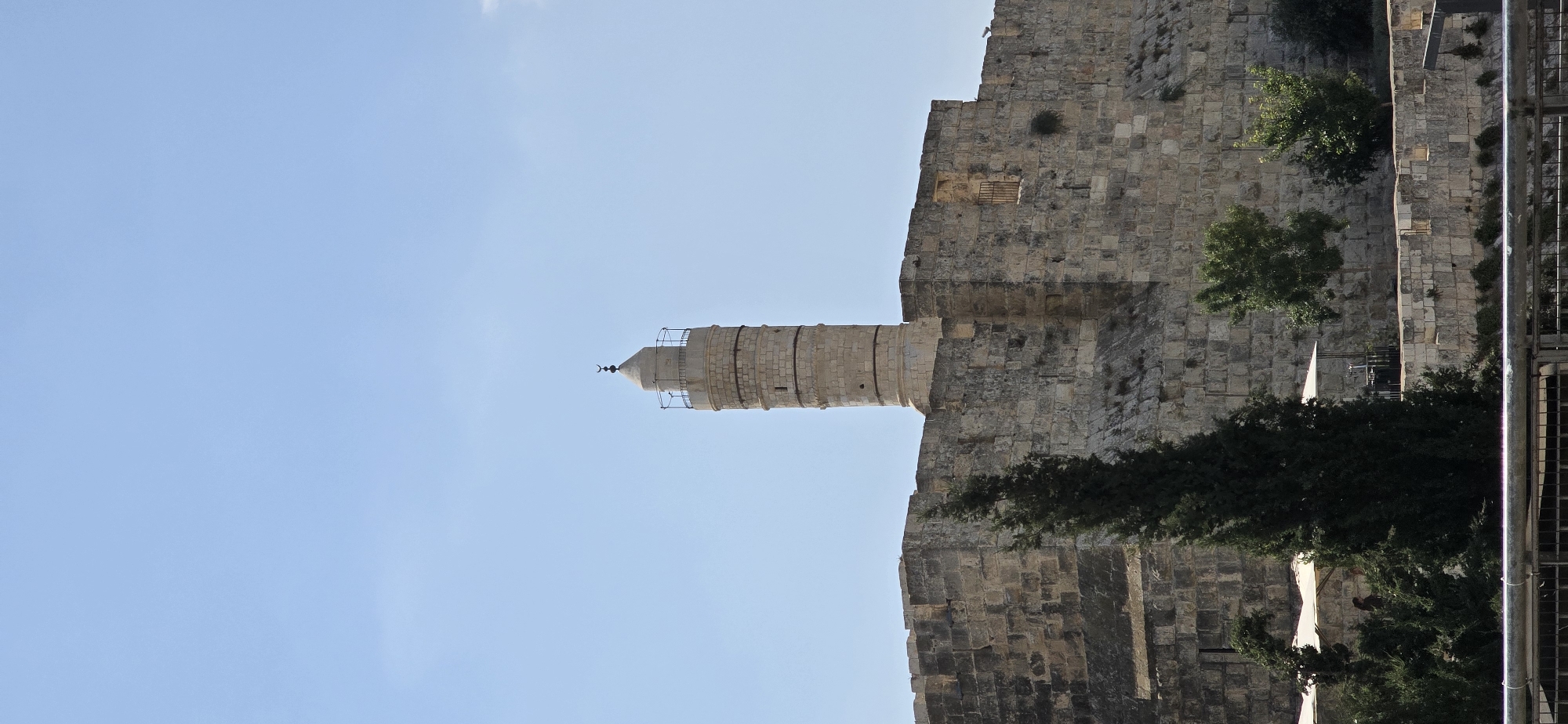 The image shows a stone minaret of a mosque rising above ancient stone fortifications under a clear blue sky in Jerusalem, Israel. The minaret is cylindrical with a conical top and a crescent moon finial. The walls are constructed of large, rough-hewn stone blocks, with crenellations visible along the top. Green trees and foliage are situated in front of and behind the walls. Some white awnings suggest outdoor seating or market stalls at the base of the walls. The image is taken from a slightly low angle, emphasizing the height of the structures.
