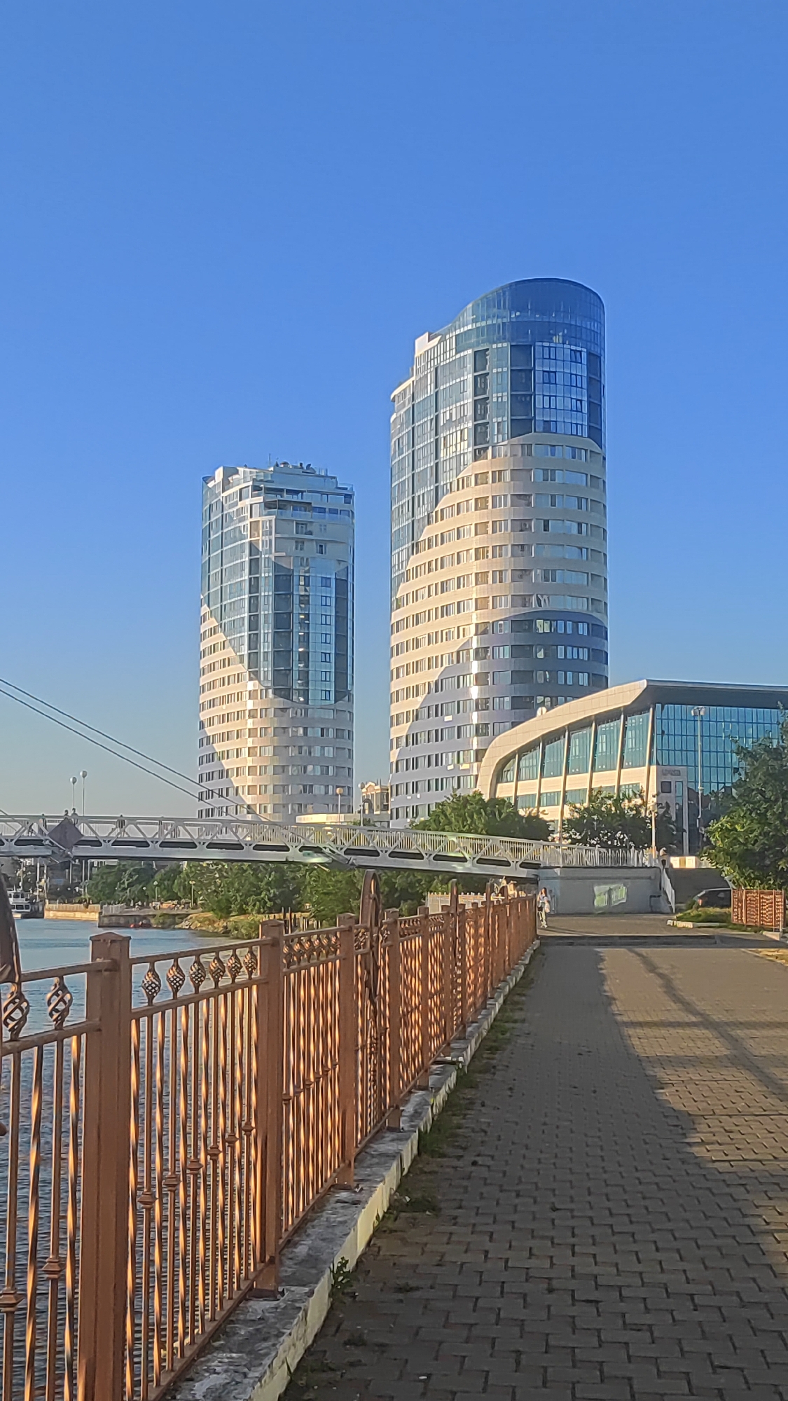 A vertical, eye-level shot captures a clear blue sky above two modern, cylindrical skyscrapers situated on the banks of a wide river, likely the Kuban River, in Krasnodar, Russia. The skyscrapers, one taller and more curved than the other, are clad in glass and light-colored panels, reflecting the sunlight. A pedestrian bridge spans the river in the middle ground, with a modern, curved building to the right of the skyscrapers. A paved walkway lines the river, bordered by an ornate, rusty-brown metal fence in the foreground. The walkway is sparsely populated by a few pedestrians. The lighting suggests it is late afternoon or early morning, casting long shadows across the pavement.