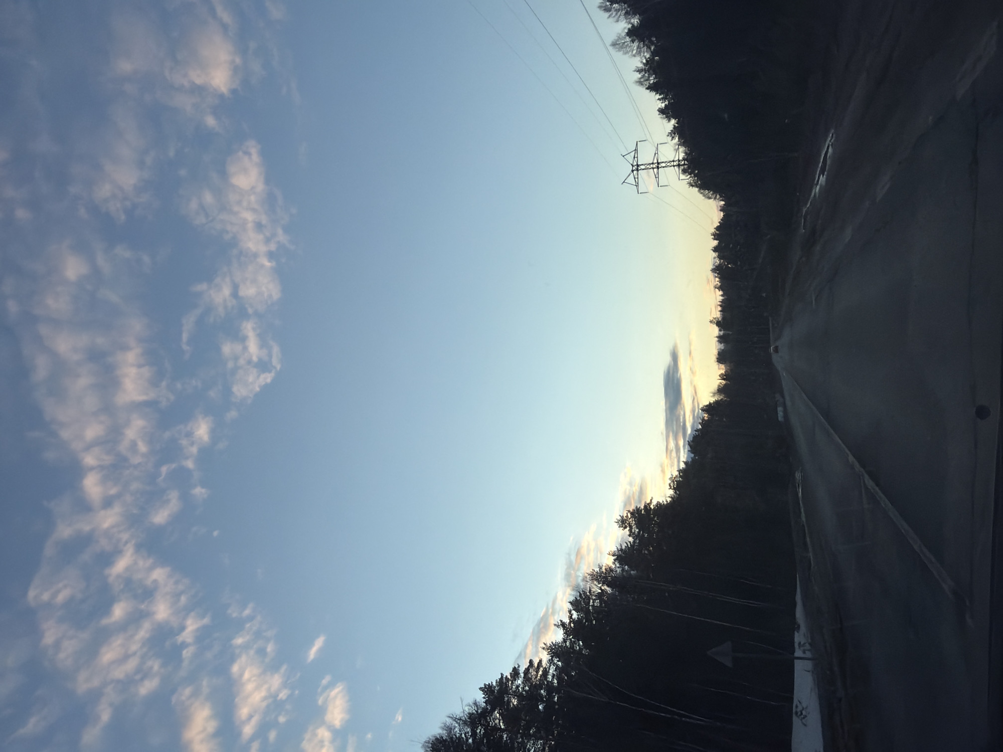 The image depicts a road stretching into the distance, flanked by a forest on one side and a cloudy sky on the other. An electrical transmission tower is visible near the center of the frame. The location is suggested to be in or around Noviy, Russia. No people or actions are visible in the image. The primary objects are the road, trees, sky, and electrical tower. The environment appears to be twilight.