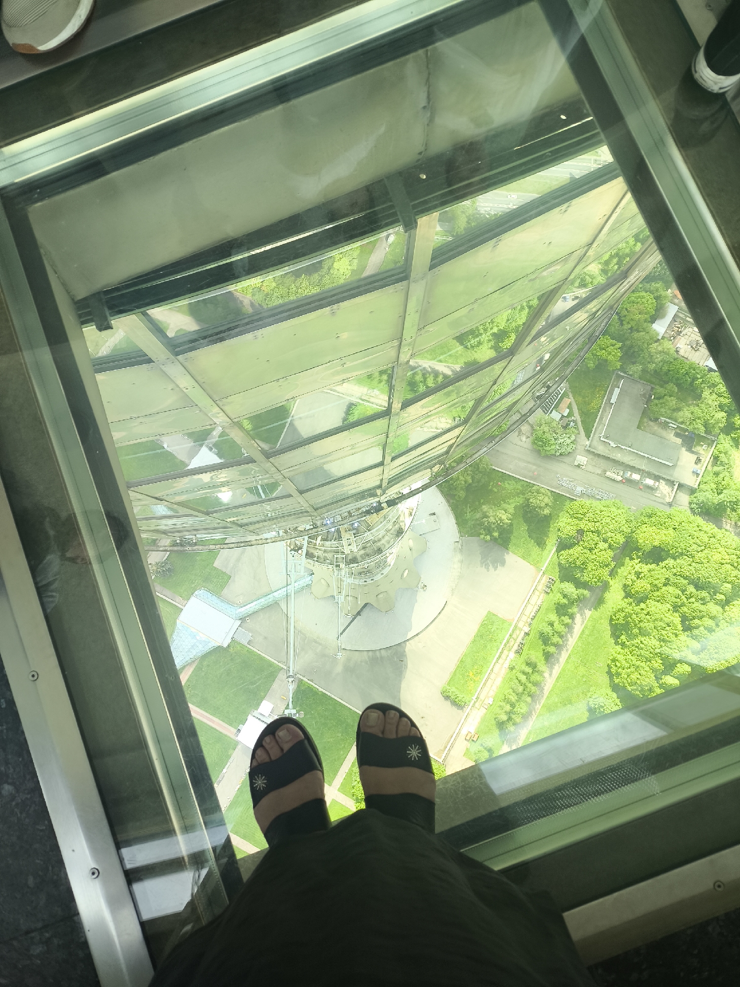 The image shows a first-person perspective of a person standing on a glass platform, likely an observation deck in a tall structure. The feet of the person are visible, wearing black sandals with a white sun or floral design. The person is also wearing a dark-colored garment, possibly a dress or skirt.
Below the glass platform, the scene depicts a view of a city with green spaces, buildings, and roads. The structure itself is visible, with steel beams and support structures descending towards the ground. The environment seems bright and sunny.
