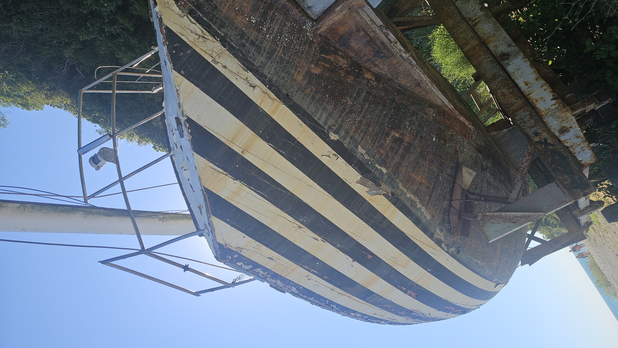 An abandoned, weathered vessel, painted with black and yellow diagonal stripes, rests on a rusty metal support structure near a body of water. The hull of the ship shows significant peeling paint and rust. A mast extends upwards, with a railing and a light fixture visible. Lush green foliage lines the background. In the lower left of the image, two people are walking on a pebble-strewn shore with the blue water of the Black Sea visible. This scene is located in Novomikhaylovsky, Russia.