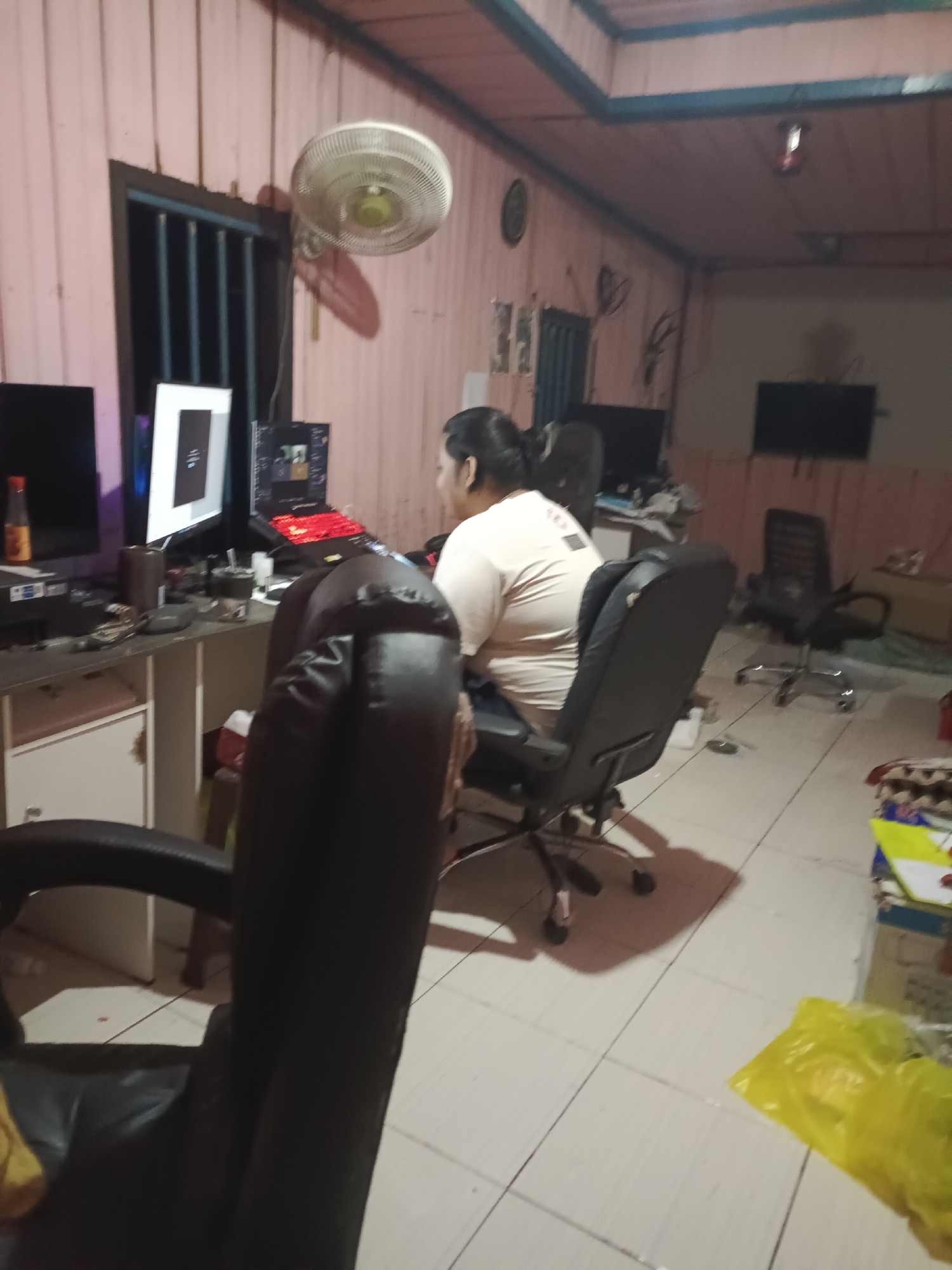 The image depicts an indoor scene, likely a room in Sangatta Selatan, Indonesia, with a person sitting at a desk in front of multiple computer monitors. The room has pink paneled walls and a tiled floor. On the desk are several computers, a keyboard with red backlighting, a mouse, and various office supplies. To the left of the desk, a bottle of sauce is visible. The person, wearing a light-colored t-shirt, is seated in a black office chair and appears to be engaged with the computers. In the foreground, another black office chair is partially visible. A ceiling fan is mounted on the wall, and a dark-colored structure, possibly a cabinet or shelf, is present further back in the room. The lighting in the room is dim, creating shadows. To the right of the desk, another office chair and a dark-colored wall-mounted screen are visible.