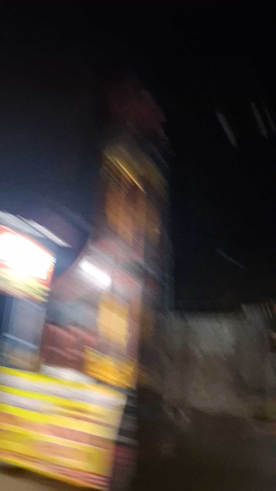 The image, captured at night in the city of Madurejo, Indonesia, is extremely blurred due to motion. In the foreground, a brightly lit structure with yellow and white horizontal stripes is visible on the left. Behind this, a red and yellow building or stall is discernible. The background is dark with some lighter, indistinct shapes and lines that might indicate other structures or lights. The overall impression is of a fast-moving scene, possibly from a vehicle, with artificial lighting illuminating parts of the street or market area.