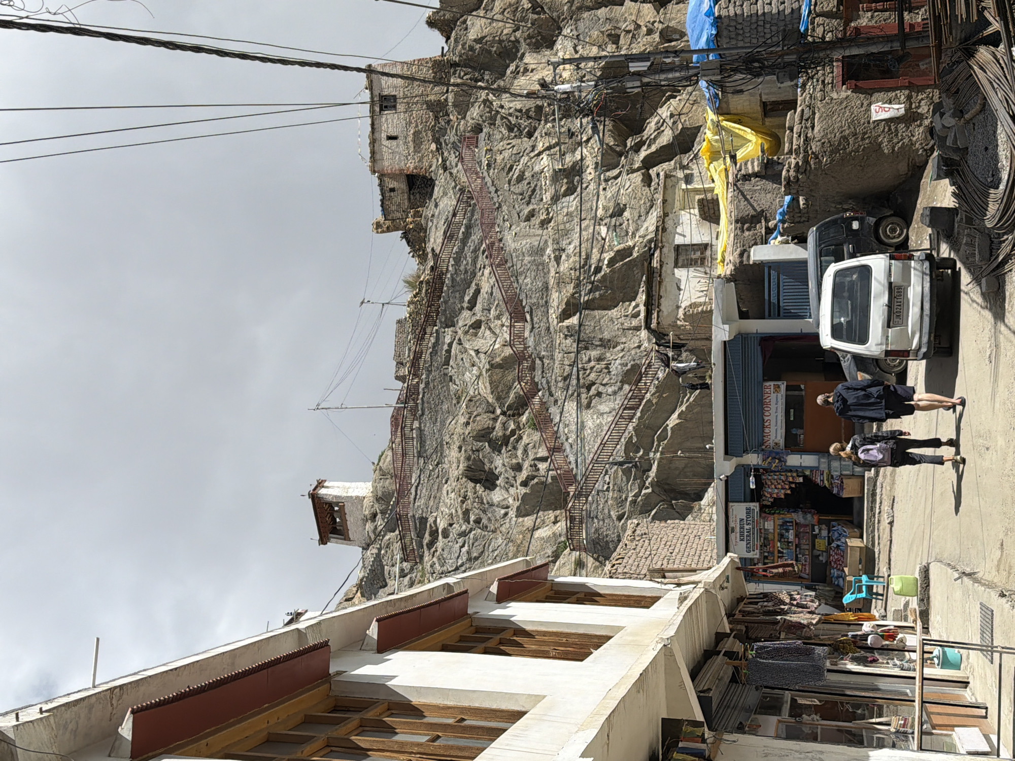 A vertical, eye-level, medium outdoor shot depicts a street in Leh, India, with a steep, rocky hillside populated by ancient structures and a long, red-railed staircase ascending to a white tower at the summit. In the foreground, a white minivan with license plate JK02AX T689 is parked, facing left, with a dark SUV behind it. Two people, a woman with a backpack and a man, walk away from the camera down the street. To the left of the street, a row of shops display goods such as clothing and bags. A bright blue plastic chair and a green barrel are visible. The street is paved with concrete, and power lines crisscross the frame. The sky is overcast.