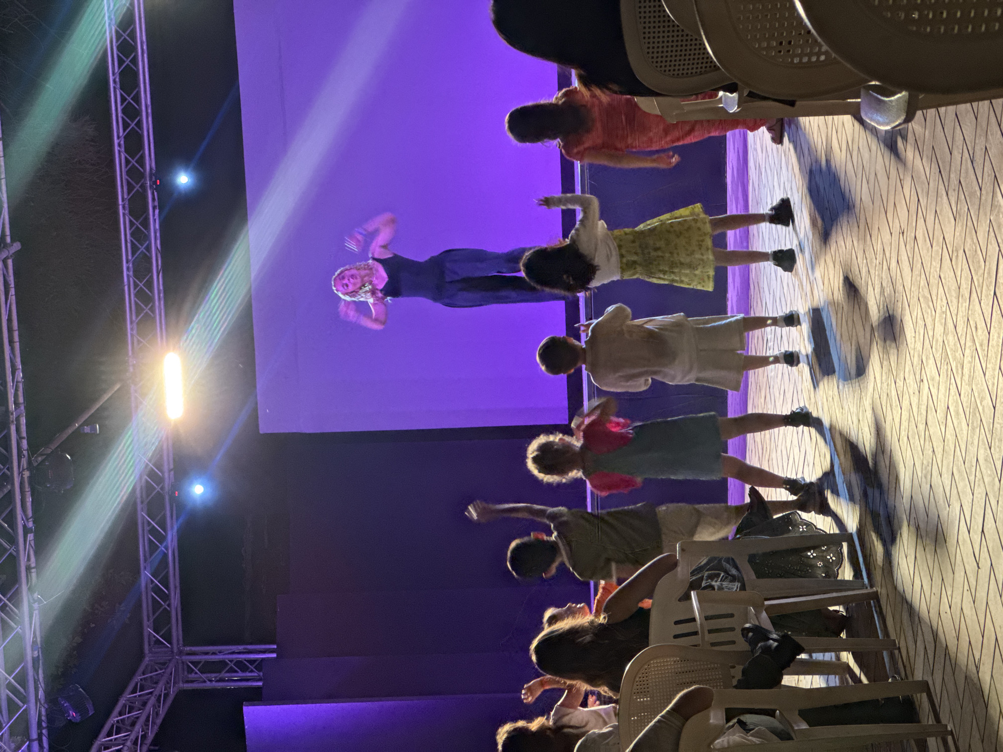 This image captures an outdoor night scene in Isola delle Femmine, Italy, depicting a performance or activity on a stage with an audience.
Centrally positioned on an elevated stage, illuminated with purple light, a blonde woman in a dark sleeveless top and dark wide-leg trousers is performing or instructing. Her arms are raised and bent at the elbows, with hands open and palms facing forward. Above the stage, a metal truss system supports multiple light fixtures, emitting bright white/yellow, blue, and green light beams into the dark sky.
In the foreground, a group of approximately five children stands on a paved surface, facing the stage. From left to right: a boy in a light-colored top and shorts stands watching; a girl in a white cardigan over a light top and a yellow floral skirt watches with her left arm slightly raised; a boy in a green t-shirt and light shorts actively raises his right arm; a girl in a pink top over a blue dress watches; and another child in a light-colored outfit is partially visible on the far right.
Behind the children, several adults are seated in white plastic chairs. One woman, wearing a dark top and a dark skirt with a patterned cut-out hem, is visible from the back. The ground is paved with light-colored rectangular tiles, intensely illuminated by the stage lighting, which casts long, sharp shadows of the children and chairs.