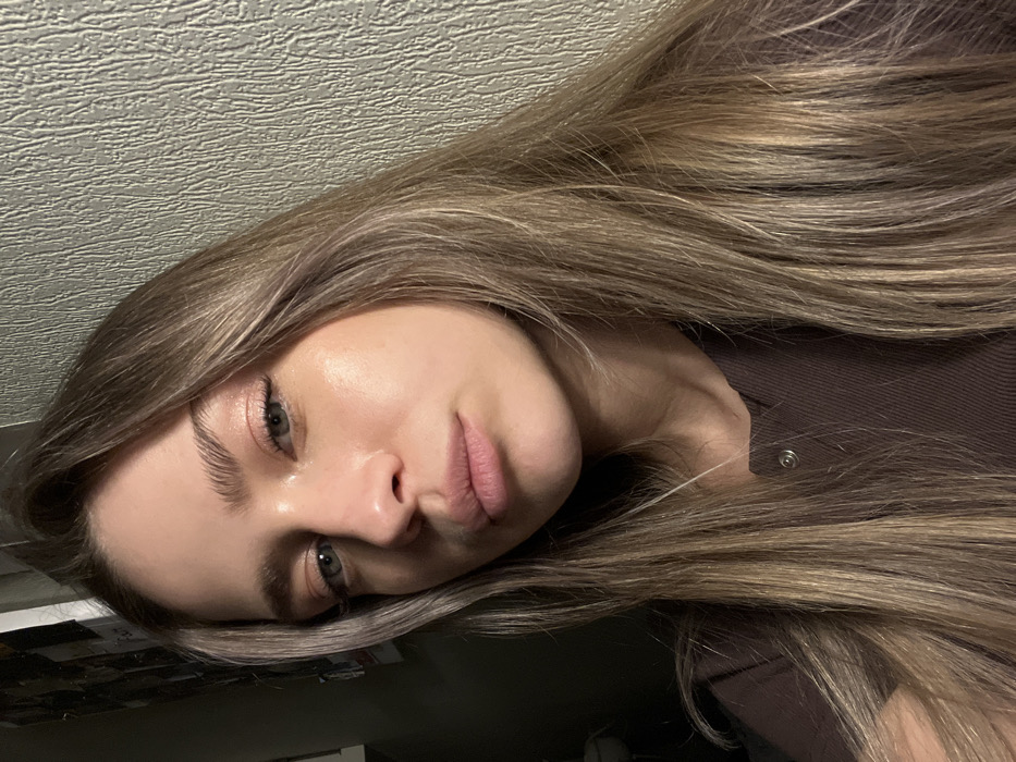 The scene is an interior setting in Saint Petersburg, Russia. A young adult female is visible in a close-up, chest-up shot. She has long, light brown hair with blonde streaks, parted slightly off-center and falling over her shoulders. Her light blue-green eyes gaze directly at the viewer, framed by full, brushed-up eyebrows. Her lips are full with a neutral to slightly pouting expression. Her complexion exhibits a subtle highlight on the forehead, cheekbones, and nose. She is dressed in a dark brown, ribbed short-sleeved top featuring a single visible silver snap fastener at the neckline. The background consists of a light-colored, vertically textured wall to her right, and a dark, indistinct area to her left, containing blurred shapes indicative of an indoor environment.