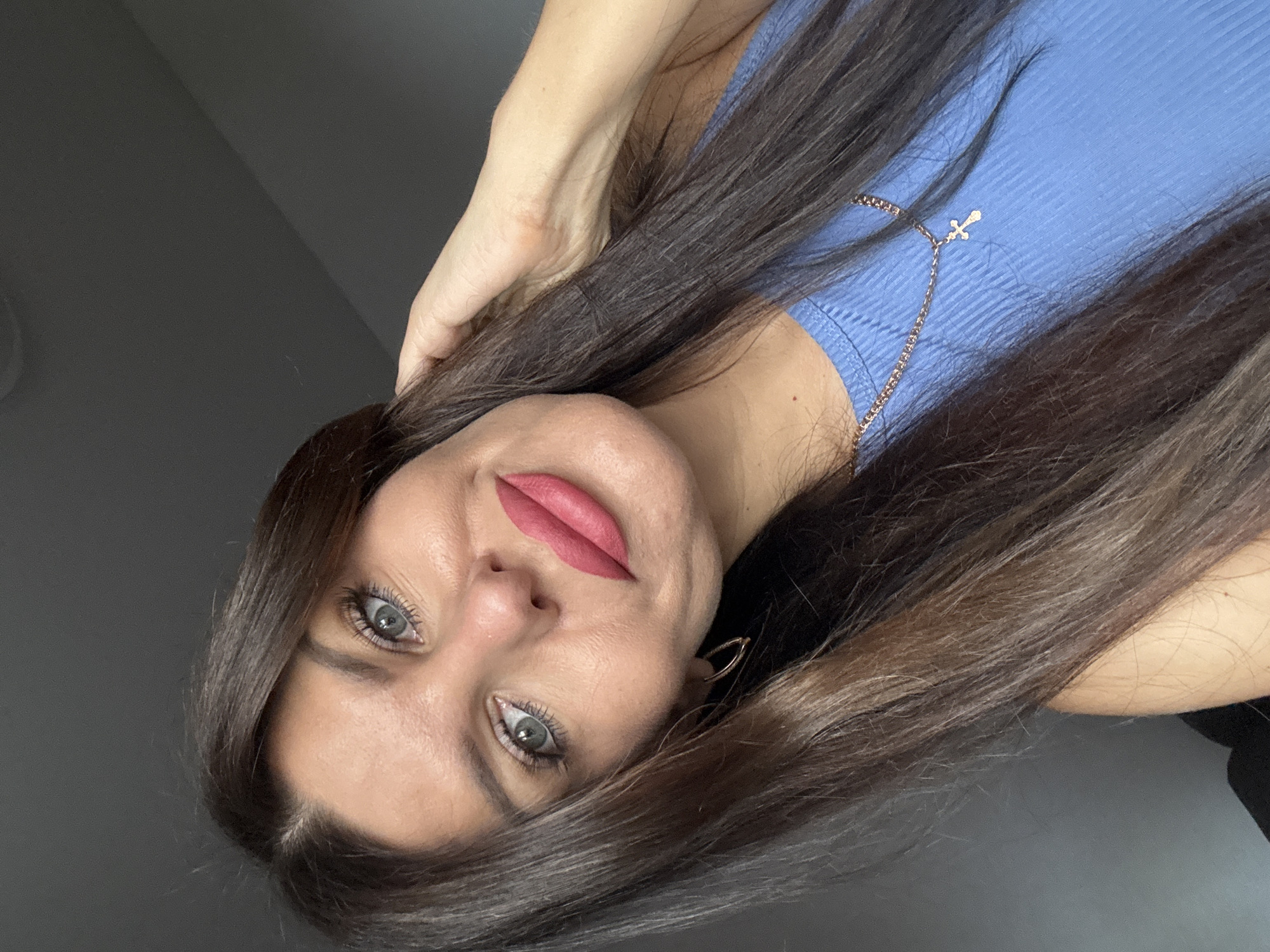 The image is a close-up, selfie-style portrait of a woman with long, dark brown hair, blue eyes, and pink lipstick. She is wearing a blue ribbed tank top and a gold chain necklace with a small cross pendant. Her right hand is raised and touching her hair. The background is a plain gray wall. The image does not contain any information that would allow for the determination of its location in Chaikovsky, Russia, or any other specific location.