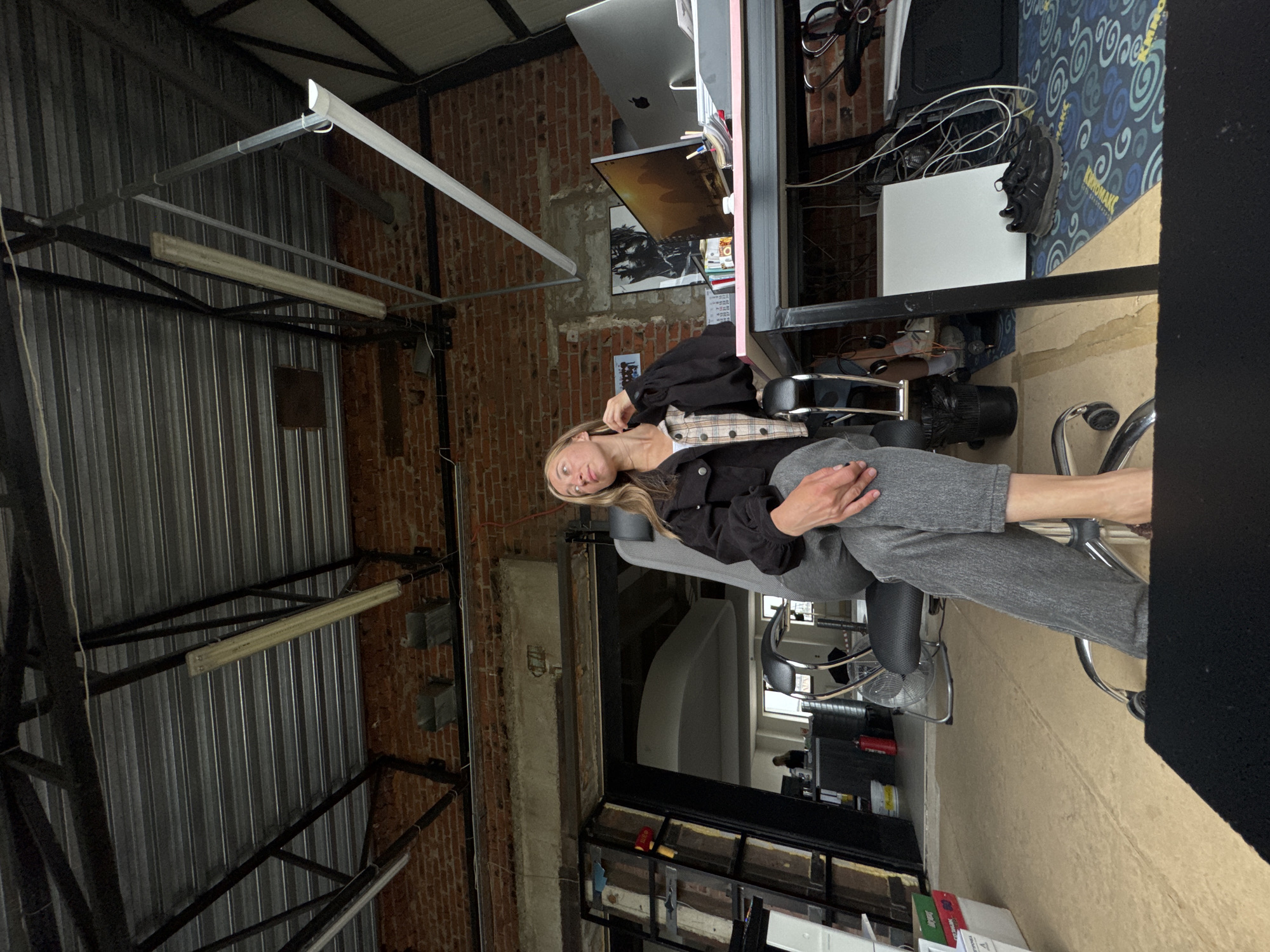 A woman with blonde hair is seated in an office chair in a loft-style workspace. She is wearing a black jacket over a plaid shirt and grey pants, with one leg extended forward. The office has exposed brick walls, a corrugated metal ceiling with industrial lighting, and a large window in the background revealing another workspace. A desk with computers and office supplies is visible to the right of the woman. The flooring appears to be a light-colored wood or concrete. The image was captured on July 18, 2023, at 12:57 PM in Chaikovsky, Russia.