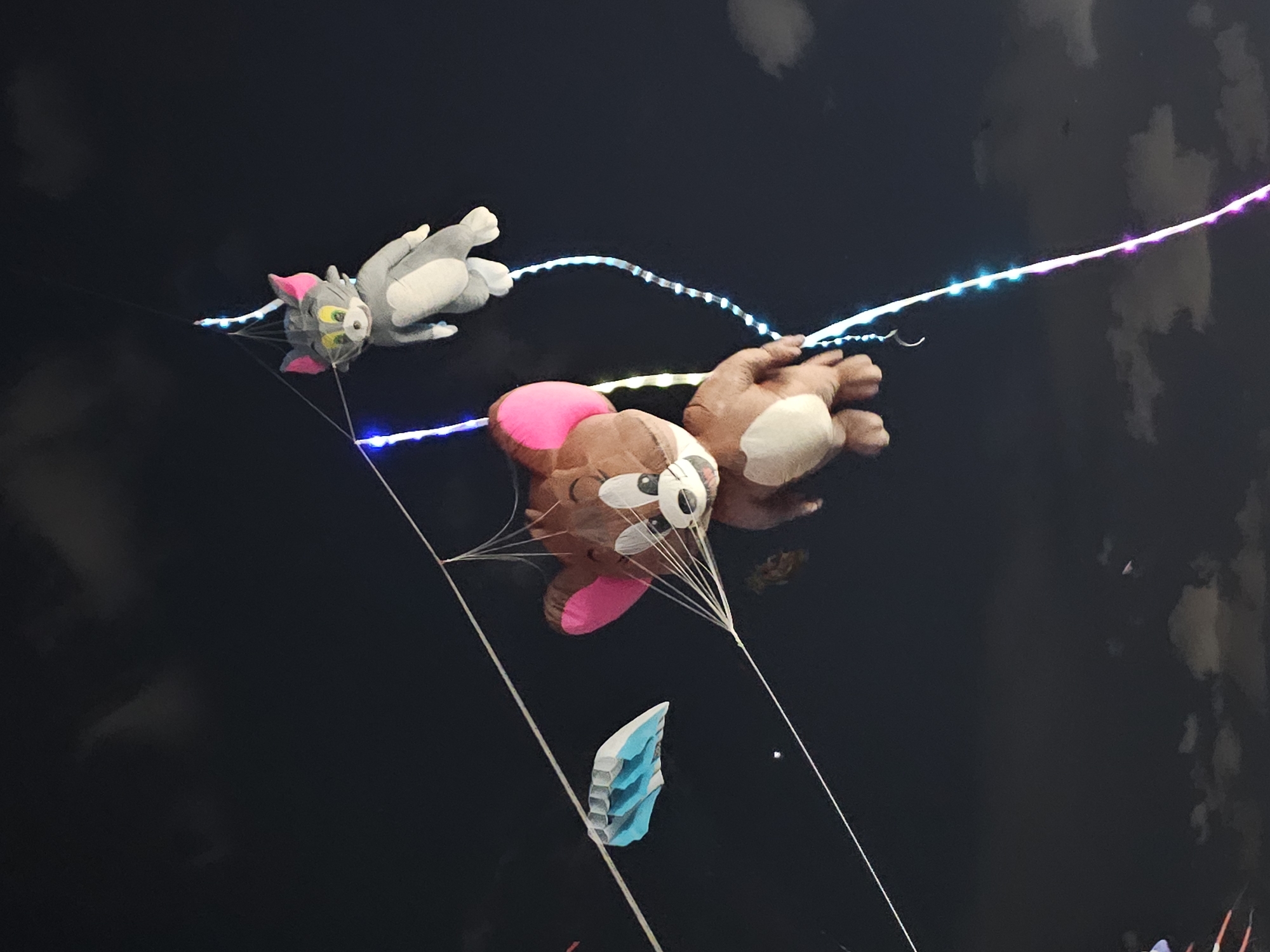 The image depicts two plush toy characters, Jerry the mouse and Tom the cat, suspended against a dark, cloudy sky. Jerry, a brown mouse with large ears and a wide smile, is larger and positioned lower in the frame. He appears to be made of fabric and stuffed. Tom, a grey cat with pink ears and a mischievous expression, is smaller and positioned higher, suspended above Jerry. Both characters are connected by strings and possibly a kite frame, suggesting they are part of a kite display. A small, blue and white kite is visible to the left of Jerry. The scene is illuminated by strings of colorful LED lights that wind around the suspension lines, creating a festive and possibly nighttime atmosphere. The cloudy sky indicates an outdoor setting, likely at night, given the artificial lighting. The presence of kites and colorful lights suggests a possible festival or event. The provided metadata indicates the image was taken in Denpasar, Indonesia.