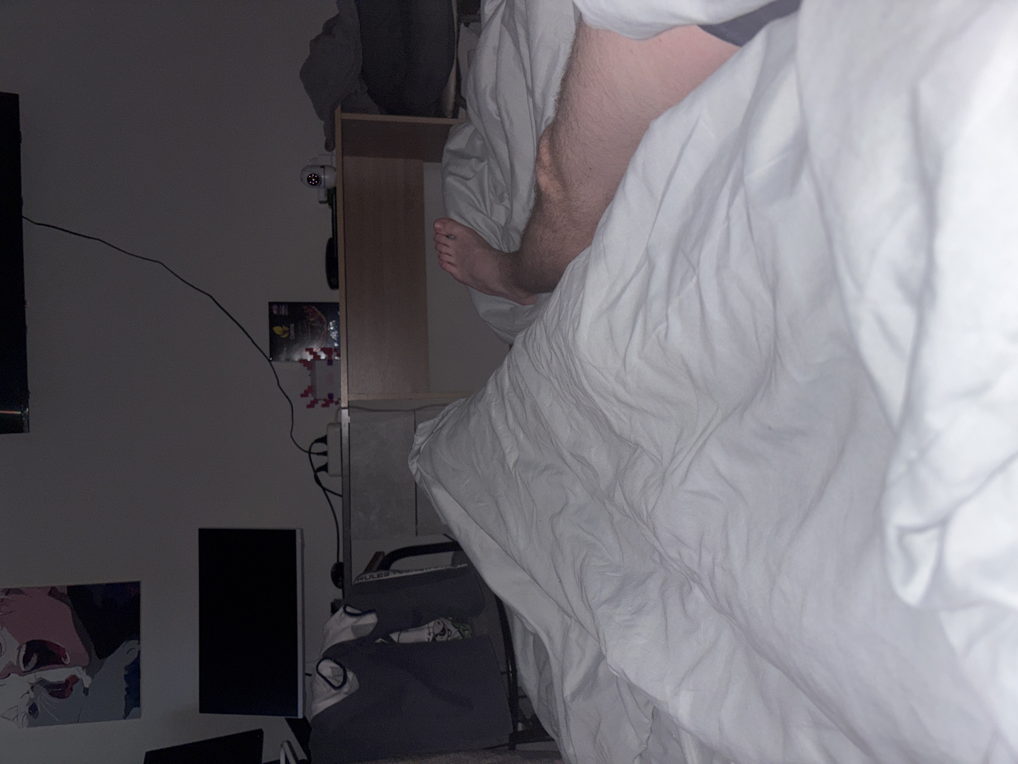The image captures an indoor scene, likely a bedroom, from a low-angle perspective and rotated counter-clockwise. The primary subject is a person's bare leg and foot, with light skin and visible hair, extending from beneath rumpled white bedsheets or a duvet. This suggests the person is resting or sleeping in bed.
The room appears dimly lit, indicating nighttime or low light conditions, highlighted by the active red infrared lights on a white security camera. This camera is mounted on the wall or a wooden shelf unit located to the right of the bed. Wires run along the wall, connecting to various electronic devices.
On the left side of the frame, a dark screen or monitor is visible. Below the camera, a poster with a yellow background and partially visible text, possibly "CRUNCH" or "CRUNCHY," is affixed to the wall, with some red and white pixel art beneath it. Further to the left and lower, another piece of artwork or a poster depicts an animated-style face with an open mouth.
Near the floor, a dark, upright rectangular object, possibly a gaming console, is positioned next to a grey fabric storage bin or laundry basket containing light-colored items, one of which has "RULES" possibly printed on it. The wooden shelf unit next to the bed holds various miscellaneous items. The overall impression is a personal and lived-in space. While the location context is provided as Sinyavino, Russia, no specific visual cues within the image confirm this geographical detail.