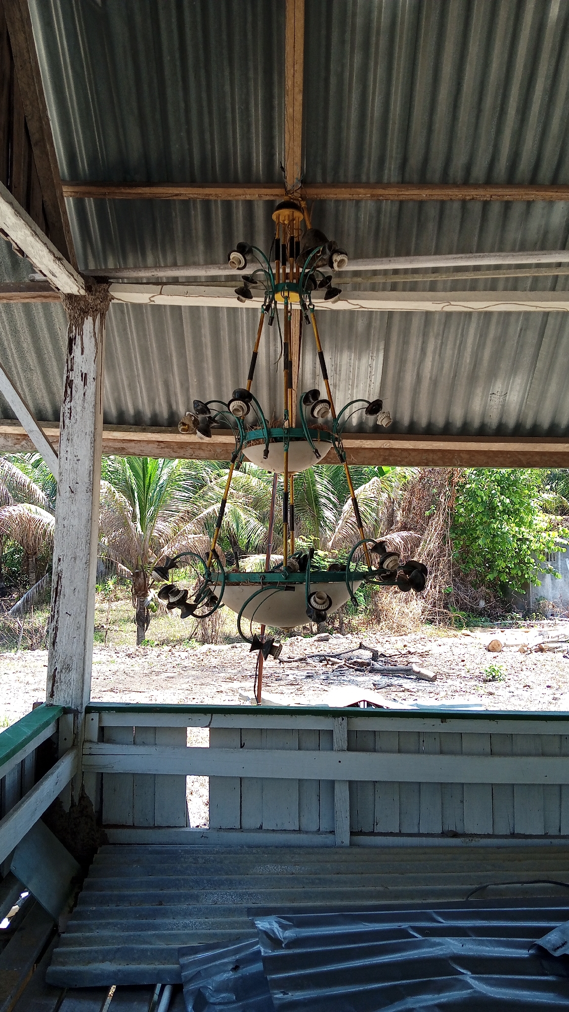 A close-up, eye-level, vertical shot captures a chandelier hanging from a corrugated metal roof supported by wooden beams in Uteunreutoh, Indonesia. The chandelier is old and appears to be made of metal, with several empty light sockets. The background reveals a tropical scene with palm trees and overgrown vegetation, suggesting a humid and possibly abandoned location. The foreground shows a wooden structure with a corrugated metal roof. The lighting is bright and natural, indicating daytime. There are no people or animals visible in the scene, and no actions or interactions are depicted. The overall impression is one of disuse and decay.