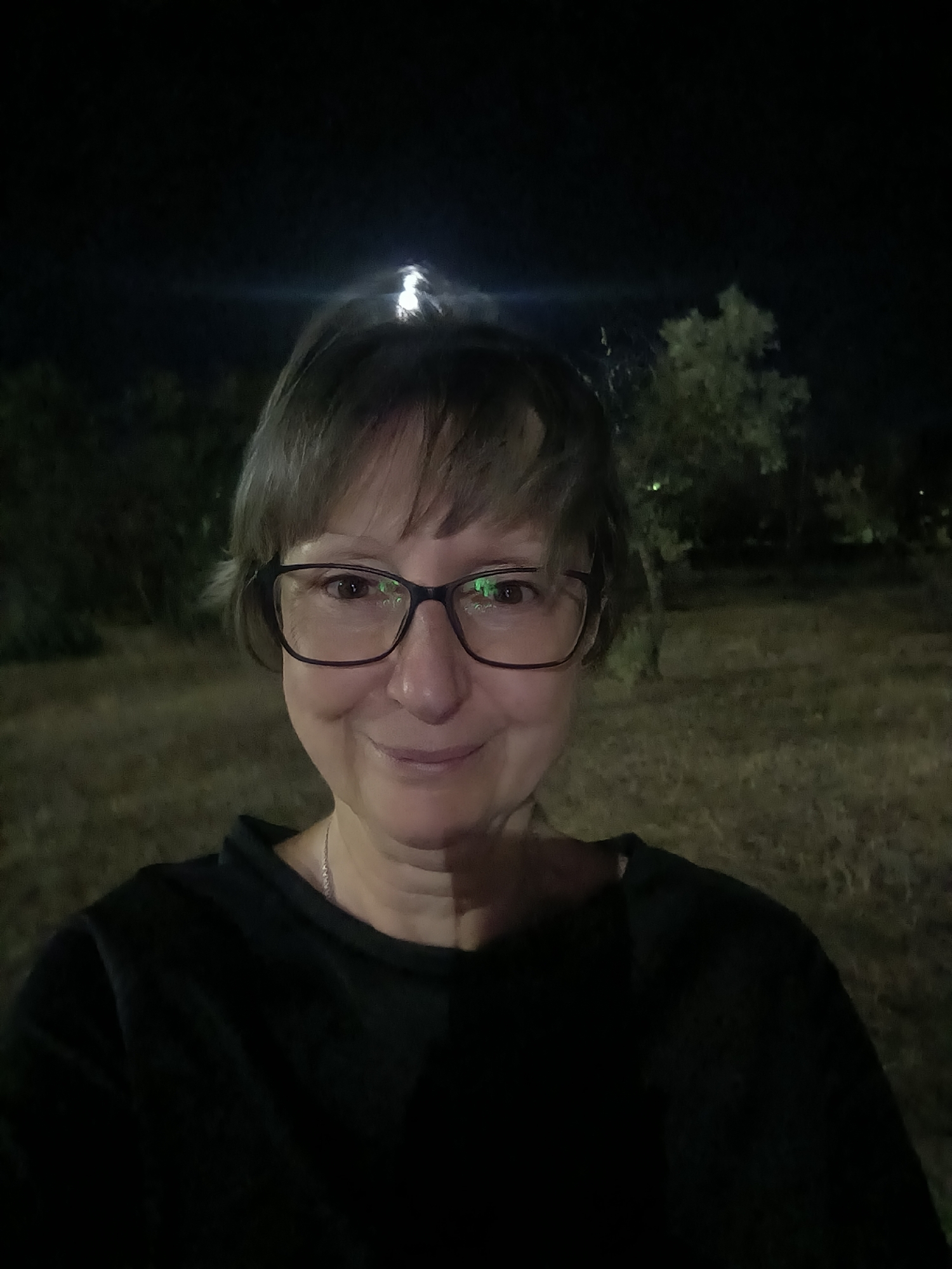 A medium shot photograph captures a woman with short, light brown hair and glasses, smiling directly at the camera. She is wearing a black crewneck shirt and a delicate silver necklace. The background is dark, with an out-of-focus grassy area and trees, suggesting an outdoor setting at night. A bright light source is visible above and behind her head, creating a lens flare effect. The overall lighting suggests artificial illumination. The location is specified as Mykolaiv, Ukraine.