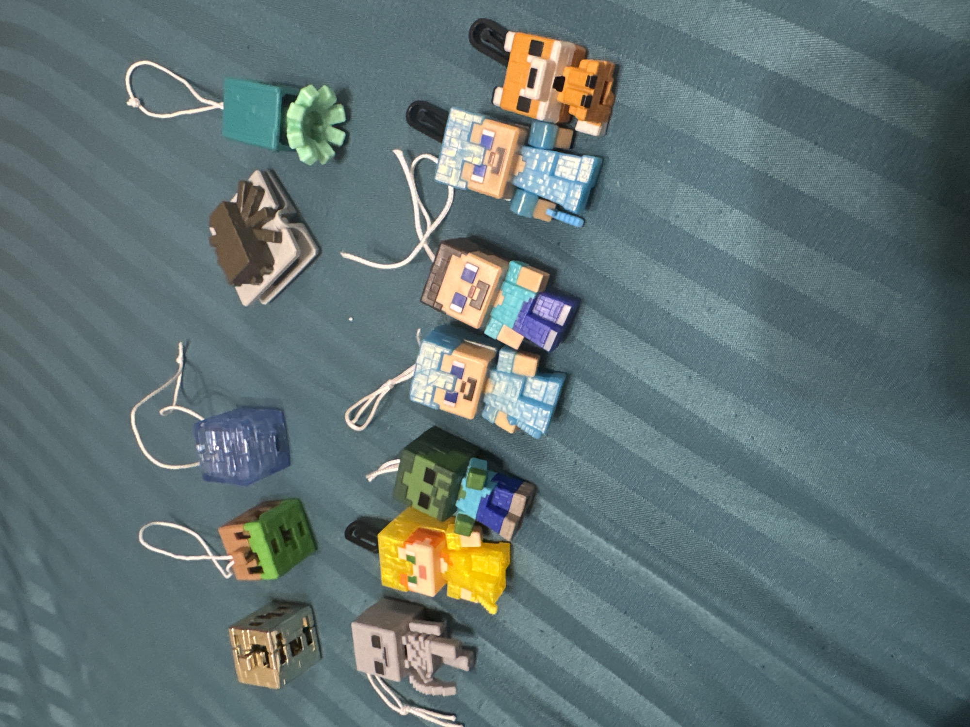 The image displays a collection of small, blocky toy figures, predominantly from the video game Minecraft, arranged on a textured, teal-blue fabric surface. Each figure is equipped with a white string loop, suggesting they are keychains or charms.
The figures include several character and creature types: an ocelot or cat, multiple versions of the player character Steve and Alex, a zombie, a piglin or villager, and a skeleton. Alongside these are block figures, resembling game elements such as a chest or block, a grass block, a translucent blue block (possibly diamond or ice), and a redstone-related block with a light green gear-like component. There is also a small brown book-like figure.
The figures are made of plastic and are colored according to their in-game counterparts. They are scattered across the fabric, which features subtle diagonal stripes. The setting appears to be indoors, likely on a bed or soft furnishing. There is no specific activity depicted; the figures are static. No time of day, weather, or emotions are discernible, and no text is visible within the image.