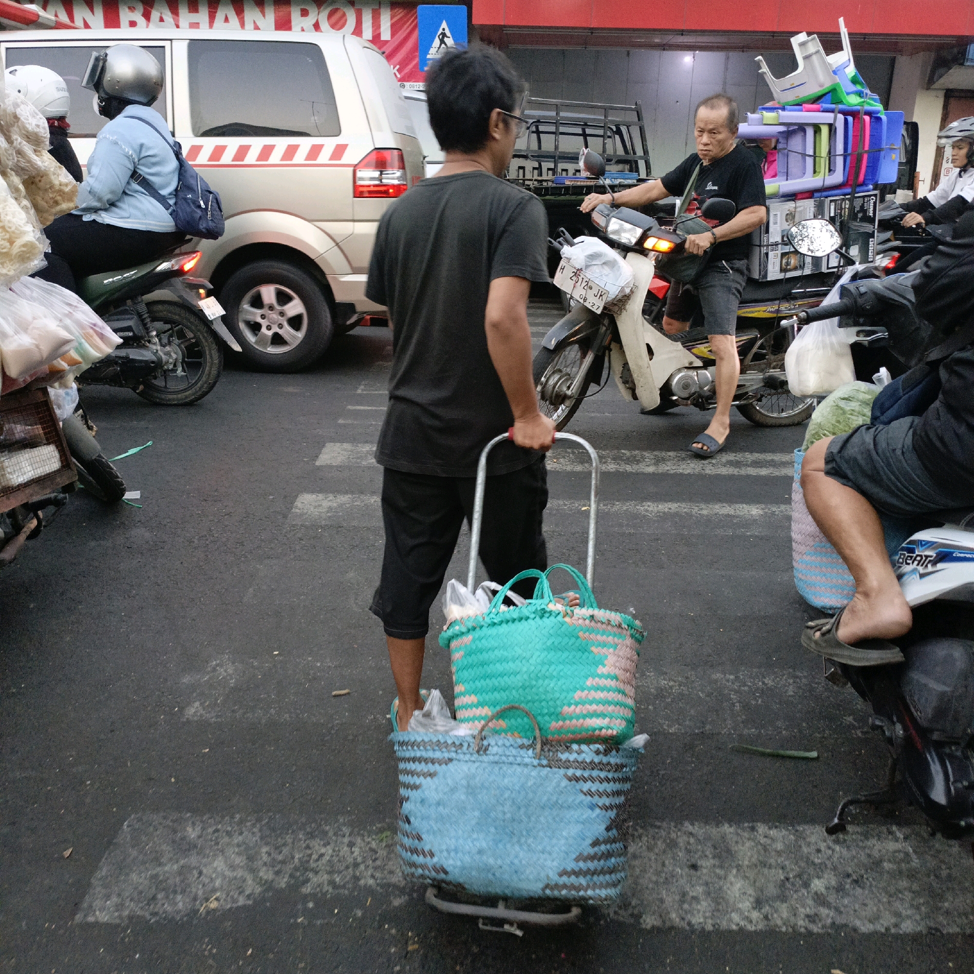 In Salatiga, Indonesia, a man wearing a black t-shirt and shorts pushes a cart with two woven baskets on a city street, likely a pedestrian crossing due to the white lines on the asphalt. The cart contains plastic-wrapped items. Behind him, several motorcycles are stopped at an intersection. One motorcycle carries a man wearing a black t-shirt and shorts, who is looking forward. Another motorcycle behind him is ridden by a person wearing a white shirt and helmet, and a third motorcycle is being ridden by someone in a black jacket. A silver van is parked to the left of the frame. A large red sign with white lettering is visible in the background. The overall scene depicts a busy urban environment with various modes of transportation.