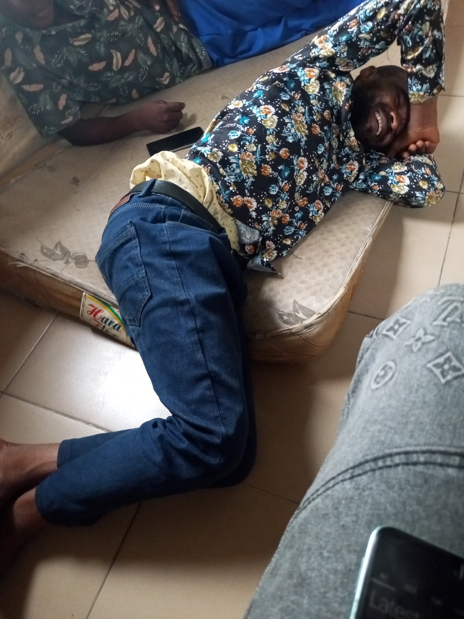 A person lies on a mattress on the floor. The person is wearing a floral print shirt and blue jeans. The person is smiling and their eyes are closed. There is a mobile phone lying on the mattress next to the person's legs. The scene appears to be indoors, possibly in a bedroom, with tiled flooring. The image quality is poor and the location in Ihalav, Nigeria is not discernible from the visual information provided.