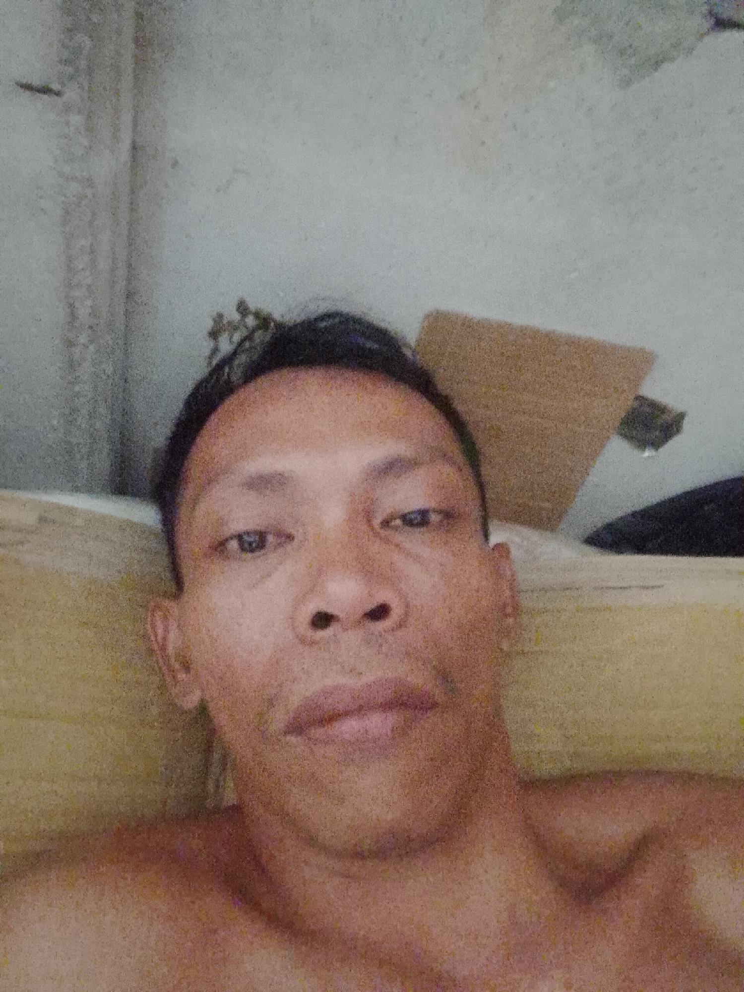 A close-up selfie of a man with dark hair and a neutral expression. He is indoors, lying down with a yellow pillow supporting his head. A piece of cardboard is visible in the background, along with a dark object. The location is presumed to be in Bukit Ketok, Indonesia, based on the request.