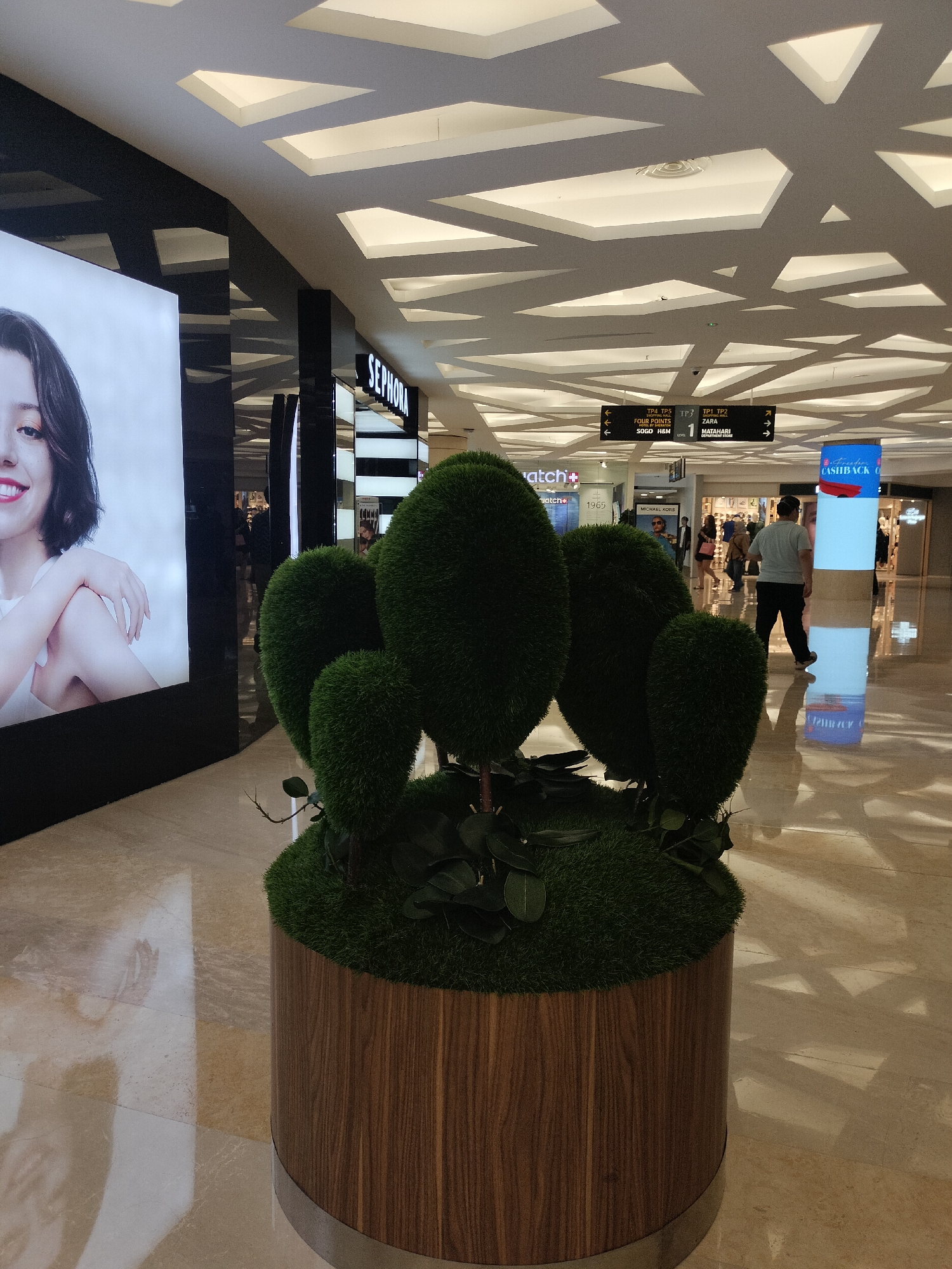 The image depicts the interior of a modern shopping mall. In the foreground, a large, round planter with a wooden exterior contains a cluster of artificial evergreen shrubs, sculpted into rounded topiary shapes. The floor is polished marble, reflecting the overhead lighting and architectural details of the ceiling, which features a geometric pattern of illuminated panels.
To the left, a large advertisement displays a close-up of a young woman's face and upper body. Behind the planter, the mall corridor extends into the background, with various storefronts visible. One prominent sign reads "SEPHORA." Further down the corridor, directional signs are mounted on the ceiling, indicating store names like "ZARA," "MATANHARI DEPARTMENT STORE," "FOUR POINTS BY SHERATON," and "SOGO H&M."
Several individuals are present in the background, walking through the mall. One person, wearing a grey t-shirt and black pants, walks away from the camera towards a tall, illuminated column displaying a "FREEDOM CASHBACK" advertisement. Another person is visible behind the Sephora store. The overall ambiance suggests a well-lit and contemporary retail environment. This scene is consistent with the interior of Tunjungan Plaza, a major shopping mall located in Surabaya, Indonesia.