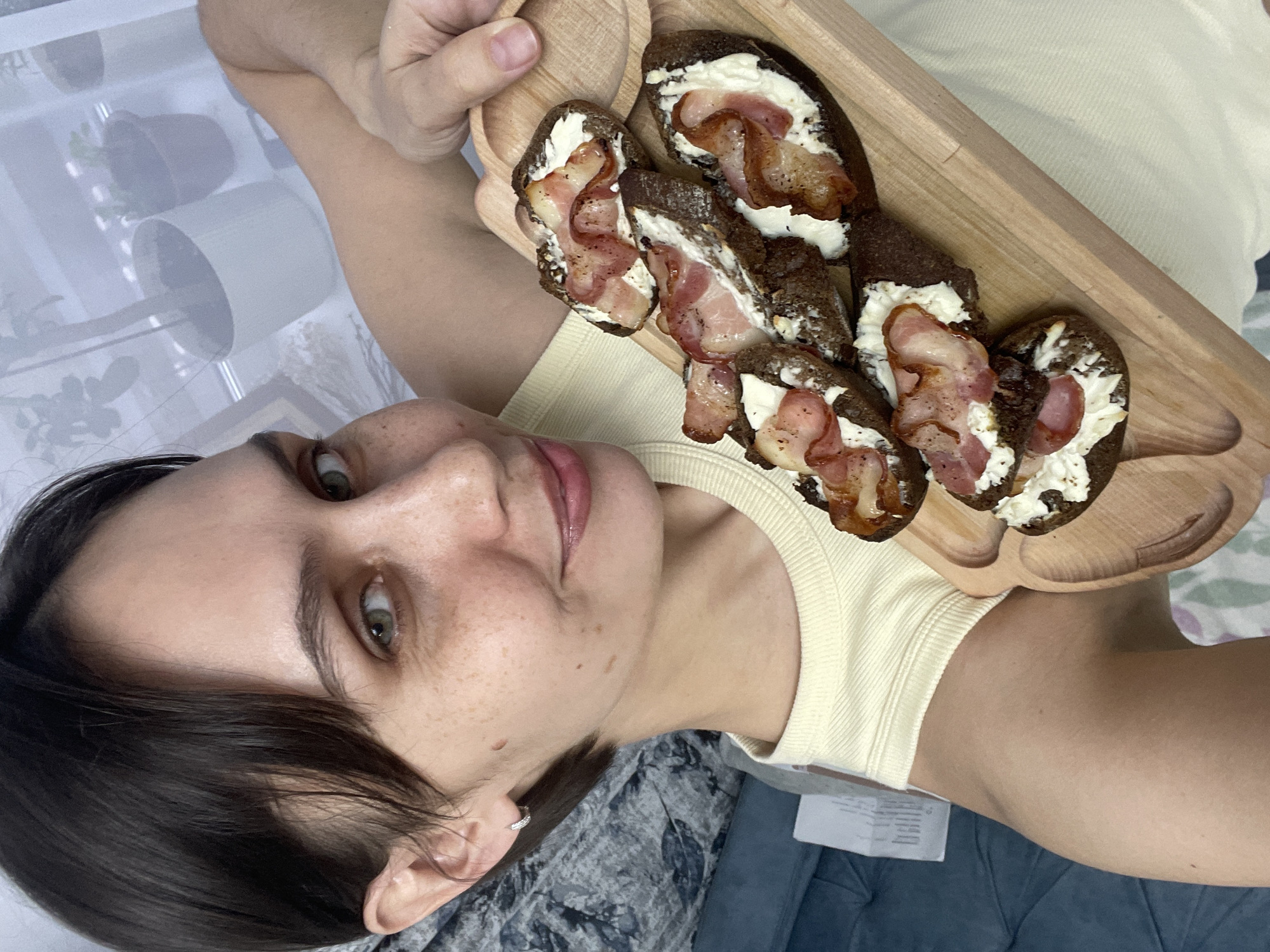 A person is holding a wooden tray with five slices of dark bread topped with a white spread and bacon. The person has dark hair and fair skin with freckles, and is wearing a light yellow top. The background includes plants and framed artwork in a partially visible room. There is no indication that the image was taken in Izhevsk, Russia.