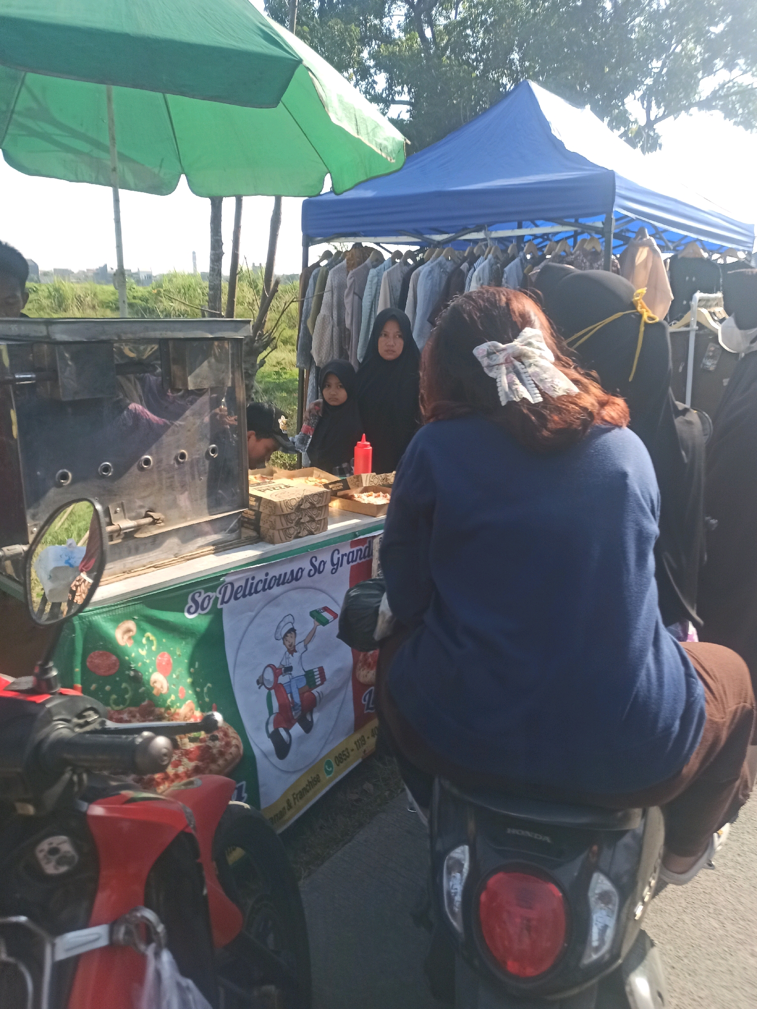 This is a medium shot, eye-level, outdoor photograph taken in Telagamurni, Indonesia. The scene depicts a street-side food stall and clothing sale under the midday sun. In the foreground, a red and black scooter is parked. A woman with long brown hair, adorned with a white and blue ribbon, is seated on the scooter, facing away from the camera and towards a food stall. The food stall features a stainless steel oven, a table displaying pizza boxes and a pizza, and a green banner with cartoon graphics of mushrooms, pepperoni, and a chef on a scooter. The banner also displays text that reads "So Deliciouso So Grand" and a phone number. Behind the food stall, a blue tent covers a rack of clothing for sale. Two young girls in hijabs are standing near the tent, and a woman in a black hijab is visible behind them. The background consists of trees and a bright, hazy sky. The ground appears to be a paved surface.