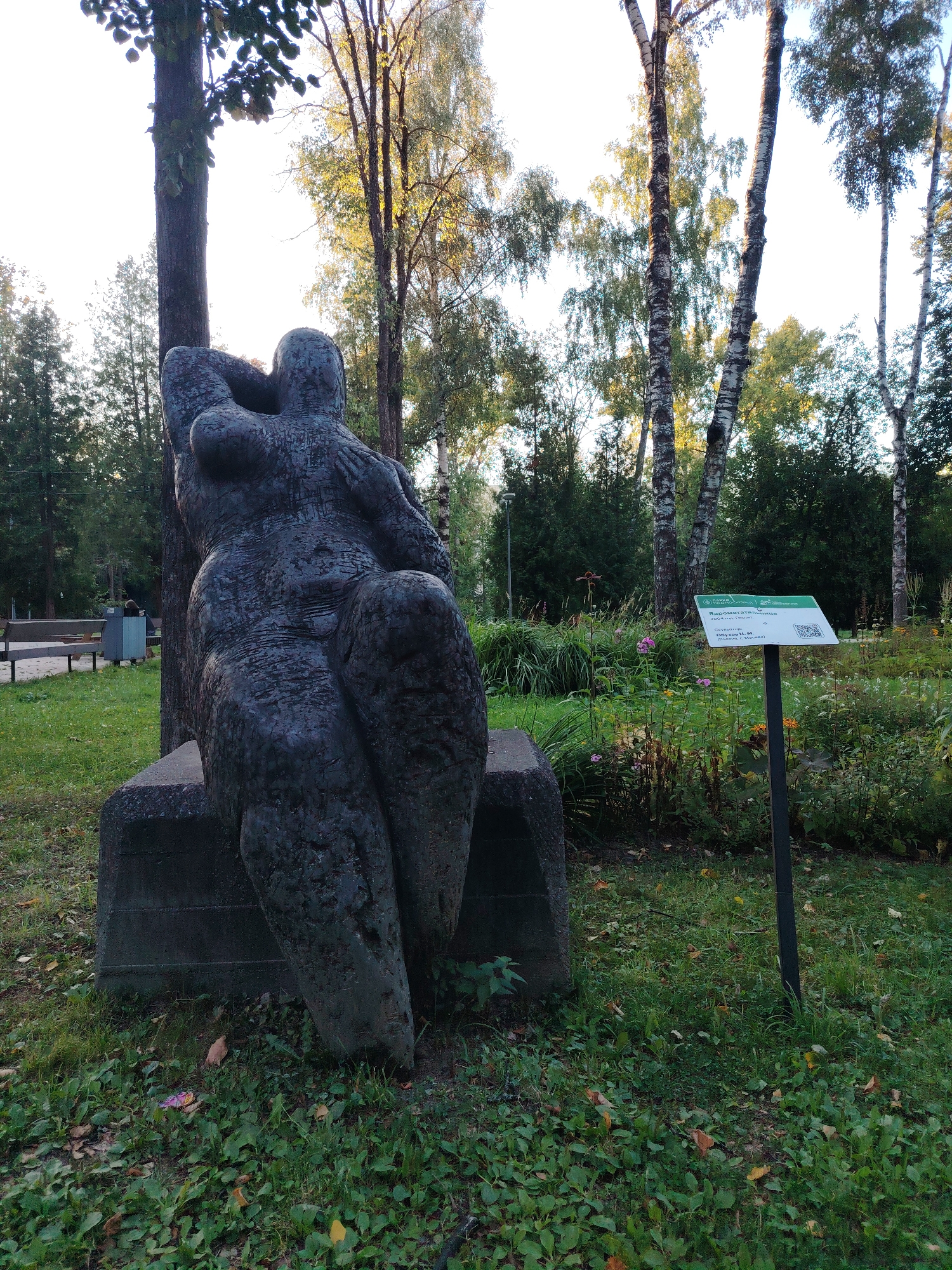 The image depicts a large, dark-colored abstract sculpture of a seated figure with rounded forms, resting on a stone base. The sculpture is situated in an outdoor park-like setting with lush green grass, fallen leaves, and a variety of trees, including birch trees with their distinctive white bark. In the background, there are more trees and dense foliage. To the right of the sculpture, a metal signpost stands with a white sign displaying text in Russian and a QR code. The sign appears to be informative, likely detailing the sculpture or its surroundings. The lighting suggests it is daytime, with sunlight filtering through the trees, creating a dappled effect on the ground. In the mid-ground, to the left of the sculpture, a wooden bench and a trash bin are visible, indicating a public recreational area. The overall scene conveys a tranquil atmosphere within a naturalistic urban park. This sculpture is likely located in the city of Solnechnogorsk, Russia, based on the provided context.