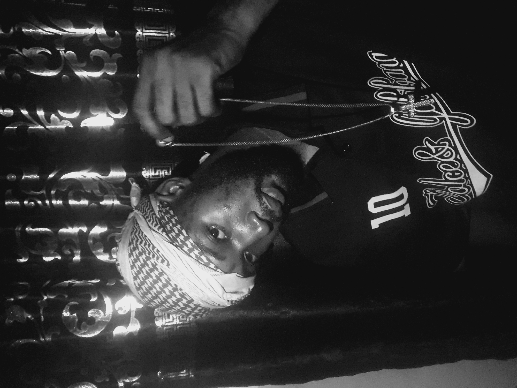 A black and white image shows a man wearing a patterned head covering and a dark shirt with the number "10" and script lettering on it. He holds a silver chain with a cross pendant. The background includes a decorative, possibly metal, surface with scrollwork patterns. The scene is dimly lit. The image's location is not identifiable as Lokoja, Nigeria from its visual elements.