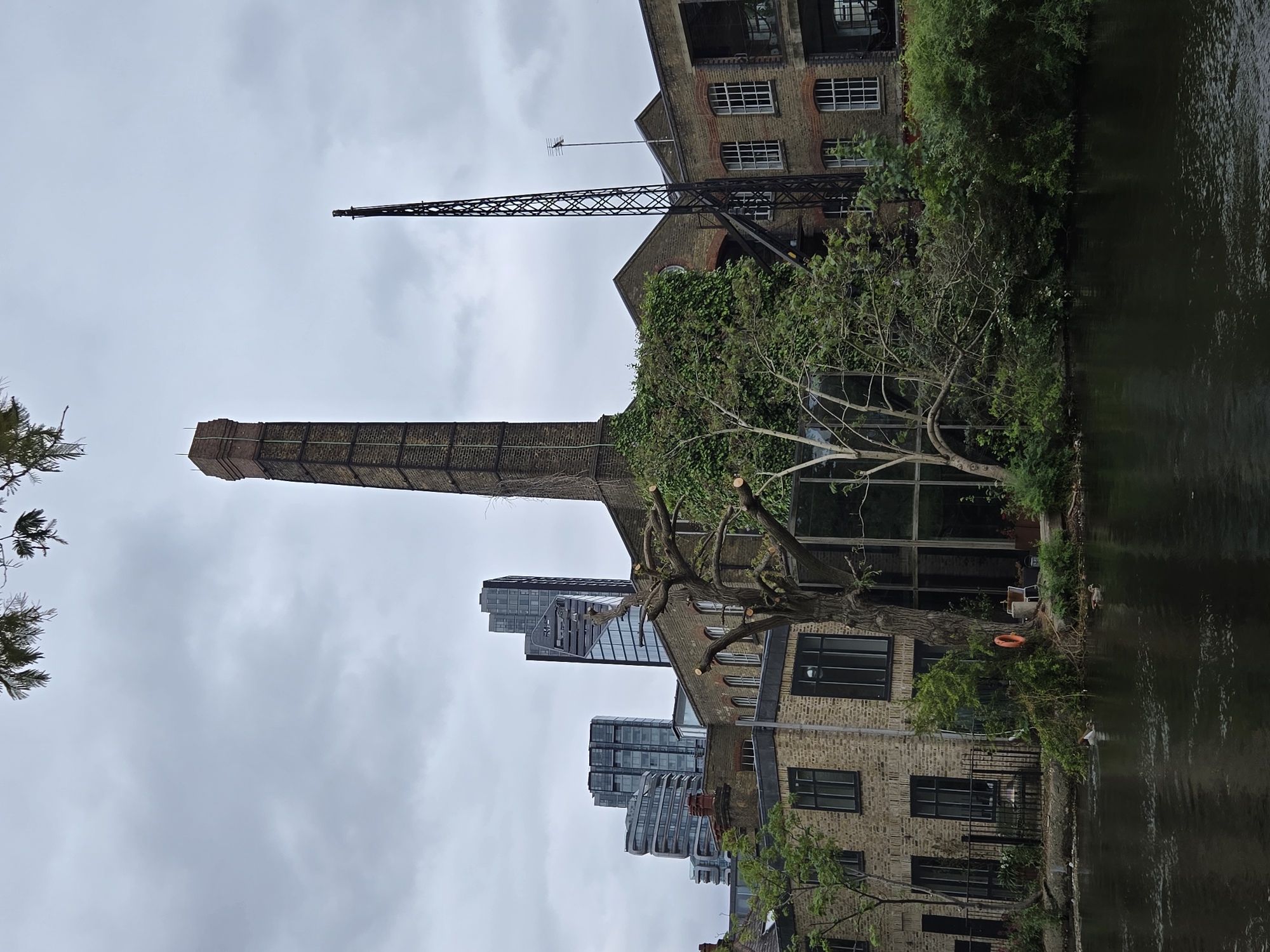 The image shows a canal scene in London, United Kingdom. A tall brick chimney stands prominently, adjacent to a brick building with windows and extensive greenery. An iron crane structure is attached to the building. Modern glass buildings are visible in the background. A bare tree stands near the water's edge, next to a building with large windows and an orange life preserver. The canal water reflects the scene. The sky is overcast. There are no people visible.