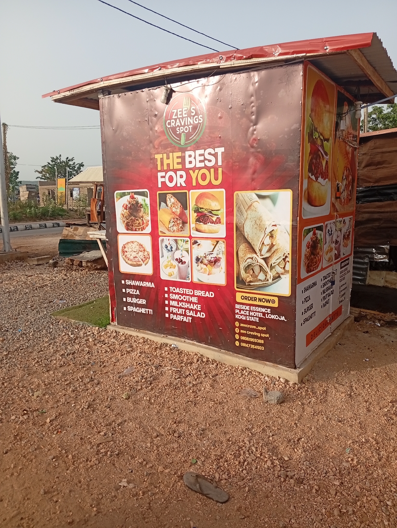 The image shows a food stand at the side of a road in what appears to be Adankolo, Nigeria. The stand, branded "Zee's Cravings Spot," is a small, enclosed structure with a red roof. The exterior is covered with advertisements for the food offered, including shawarma, pizza, burgers, spaghetti, toasted bread, smoothies, milkshakes, fruit salad, and parfait. Contact information for ordering is displayed, including a location near Essence Place Hotel in Lokoja, Kogi State, social media handles, and phone numbers. The ground around the stand is gravel and dirt. The sky is overcast and some infrastructure or other buildings are visible in the distance. A single shoe lies on the ground near the base of the food stand.
