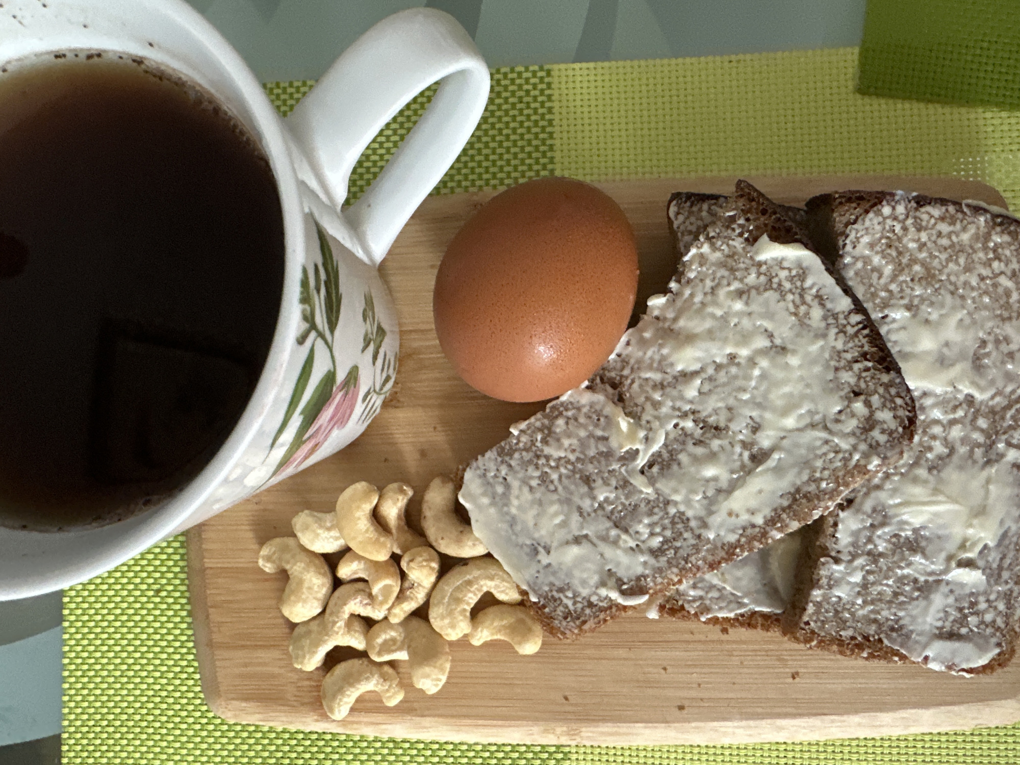A mug of dark liquid, likely coffee or tea, sits on a green placemat. Beside it on a wooden cutting board are a pile of cashews, a brown egg, and three slices of dark bread spread with a white substance, possibly butter. The mug is white with a floral design. The scene appears to be set on a table. There are no people or animals visible. The image does not contain any information that would indicate its location in Podolsk, Russia.