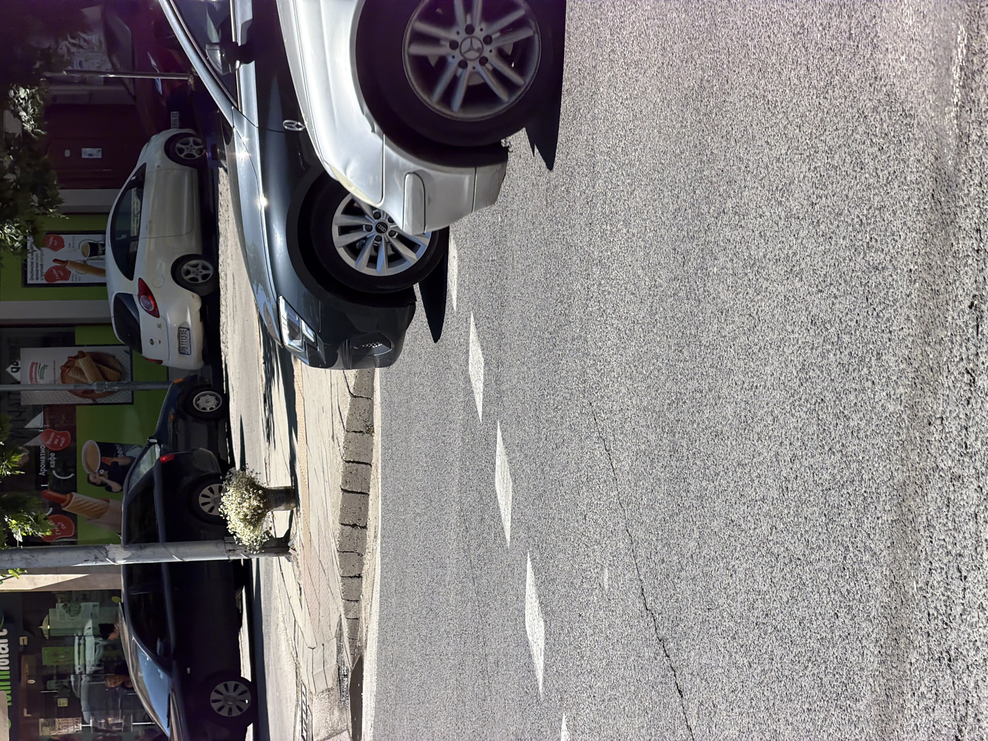 The image captures a street scene in Plovdiv, Bulgaria, featuring a row of parked cars along the sidewalk. From top to bottom, there is a white Mercedes, a black Audi, a white Ford Fiesta, and a dark sedan. The vehicles are parked adjacent to commercial buildings with advertisements and signs in Cyrillic script. A sidewalk runs alongside the buildings, and there is a flowerpot placed near the curb between the cars. The street itself is paved with asphalt and has painted white markings.
