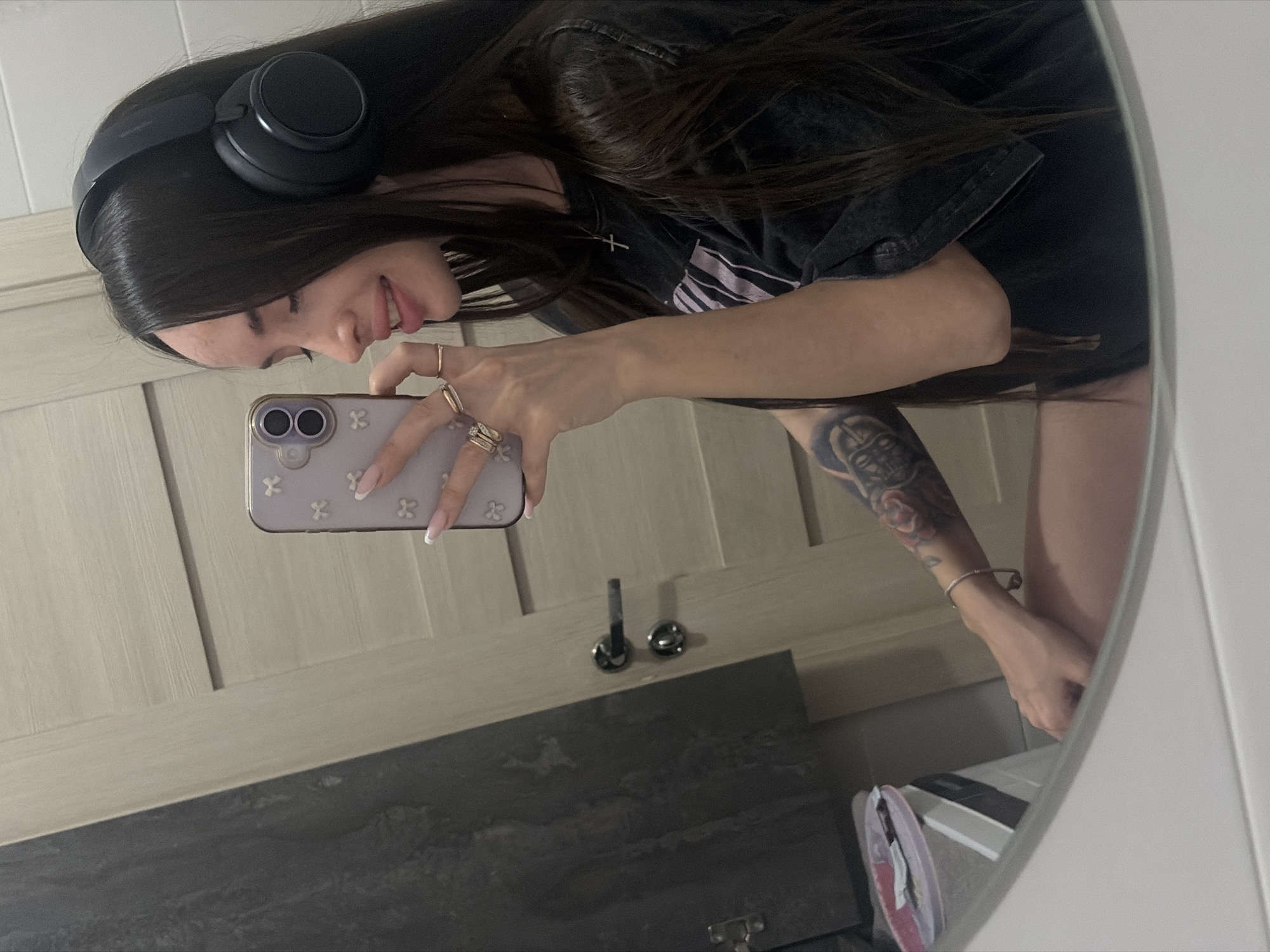 A person is taking a selfie in front of a mirror. The person has long dark hair and is wearing headphones. They are holding a phone with a light purple case decorated with white clovers. The person is wearing a dark t-shirt and a necklace with a cross pendant. They have several rings on their fingers and a tattoo on their left arm. The mirror reflects a bathroom with a light-colored wooden cabinet, a dark countertop, and a double towel rack. A glimpse of the lower part of the body shows a bare leg. The reflection is in a round mirror with a white frame. It is impossible to determine if this image was taken in Saint Petersburg, Russia.