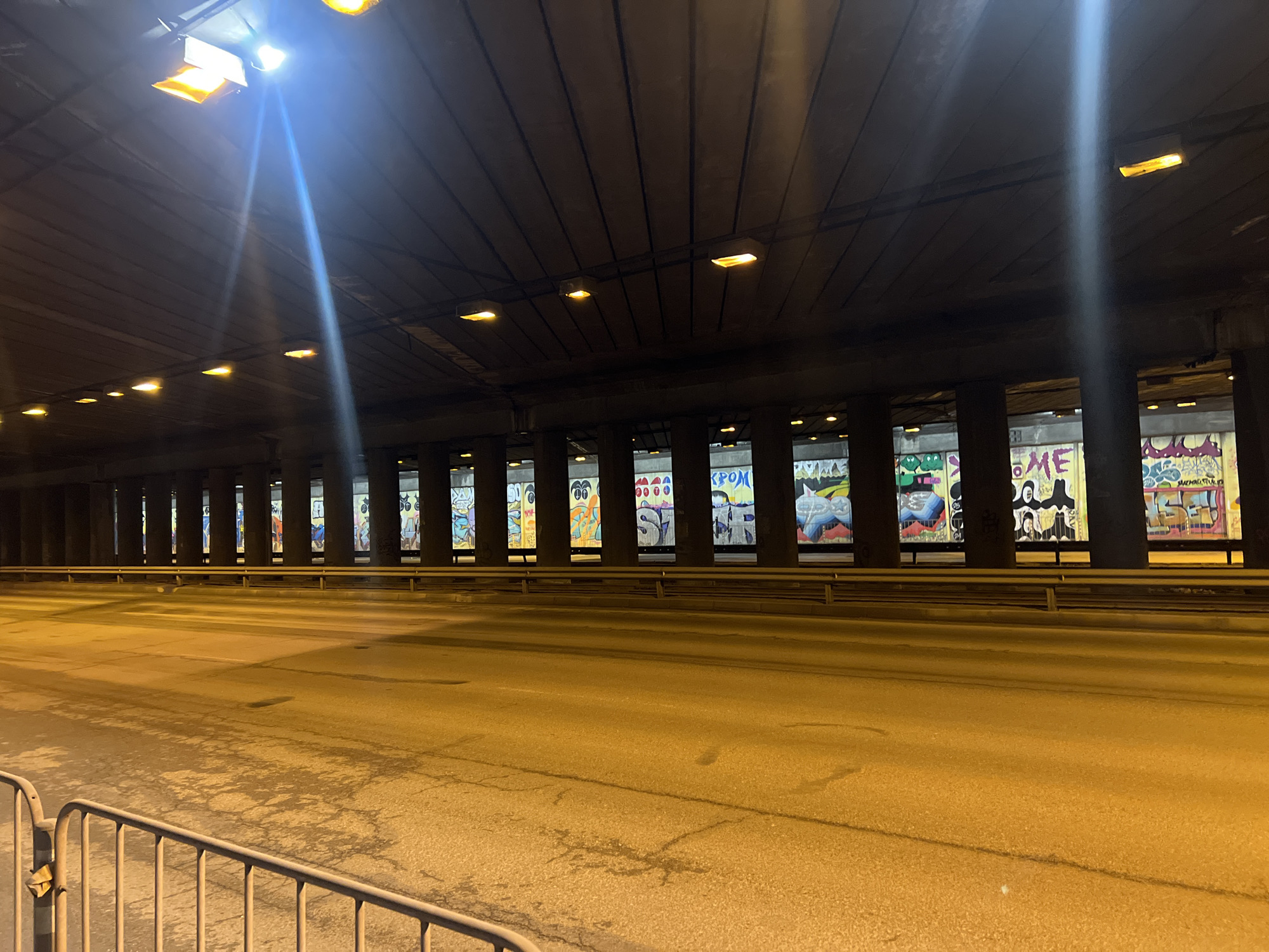 The image shows a long, empty underpass or tunnel in Sofia, Bulgaria. The road surface is asphalt, illuminated by a series of overhead lights that cast a yellow glow. A low, metal safety barrier is in the foreground. The walls of the underpass are lined with pillars separating large openings, the surfaces of which are covered with extensive graffiti art. The artwork varies in style and subject matter, covering the entire available area of each opening.