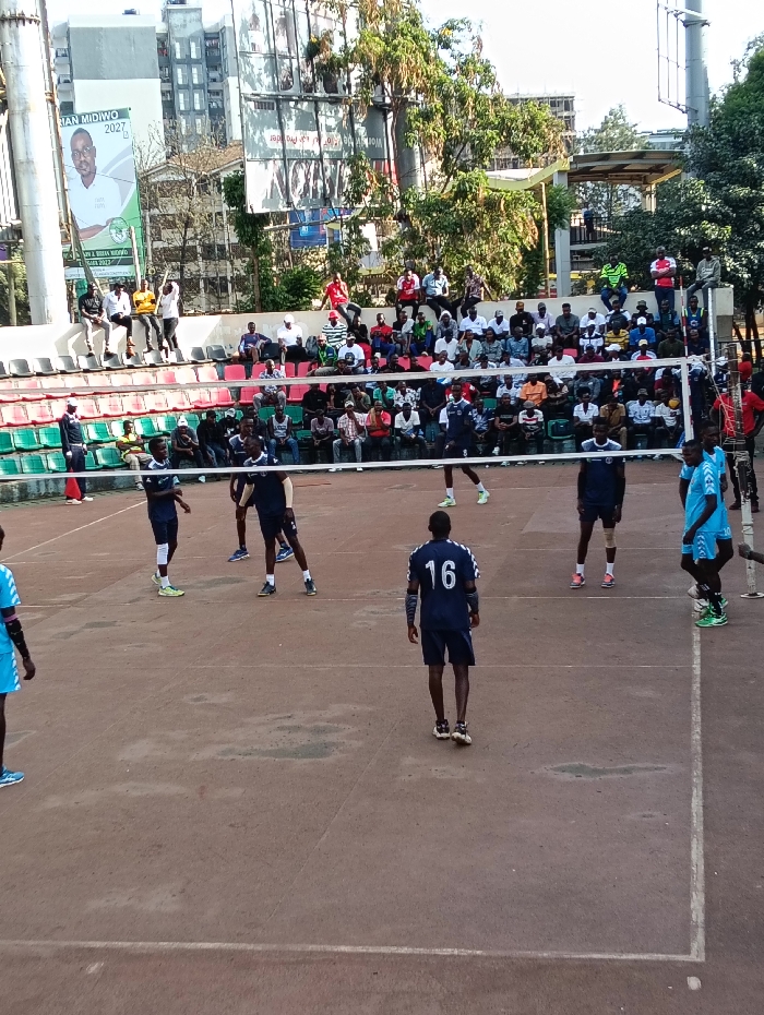 An outdoor volleyball game is depicted on a reddish-brown hard court within an urban setting in Nairobi, Kenya. Approximately eight male players are distributed across the court, wearing dark blue and light blue sports uniforms. One player, wearing a dark blue jersey with the number 16 on the back and white shoulder stripes, dark shorts, white socks, and yellow athletic shoes, is in the foreground facing away from the viewer. A white volleyball net is stretched across the court. Tiered concrete spectator stands, painted in alternating green and red sections, are visible behind the court and are populated by numerous male spectators in casual attire. In the background, large billboards are present; one prominently displays "BRIAN MIDIWO 2027" and "Your NBI MP 2027" alongside a portrait. Other urban buildings, some appearing residential or commercial, and mature trees complete the background under bright daylight conditions. The court shows visible wear, and white boundary lines are delineated.