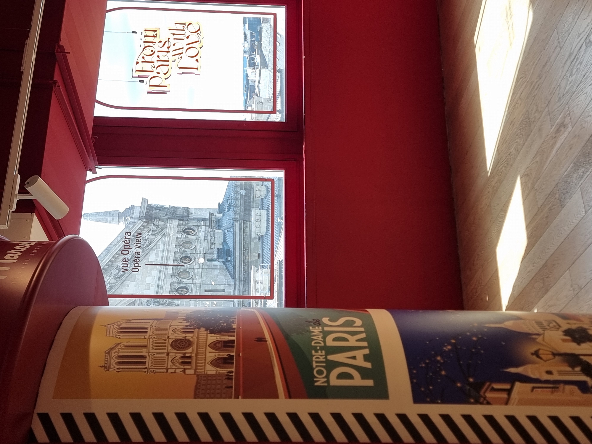 The image shows an indoor scene in Paris, France, featuring a view of the city through two adjacent windows. One window has the text "From Paris With Love" on the glass. The second window shows a section of the city, including what appears to be a cathedral and various buildings. In the foreground, there is a cylindrical object with an image of Notre Dame de Paris on it, along with the words "NOTRE-DAME de PARIS." The object has a striped pattern on the bottom. The interior walls and window frames are painted a deep red. Light from the window shines on a wooden floor in the upper-right corner.