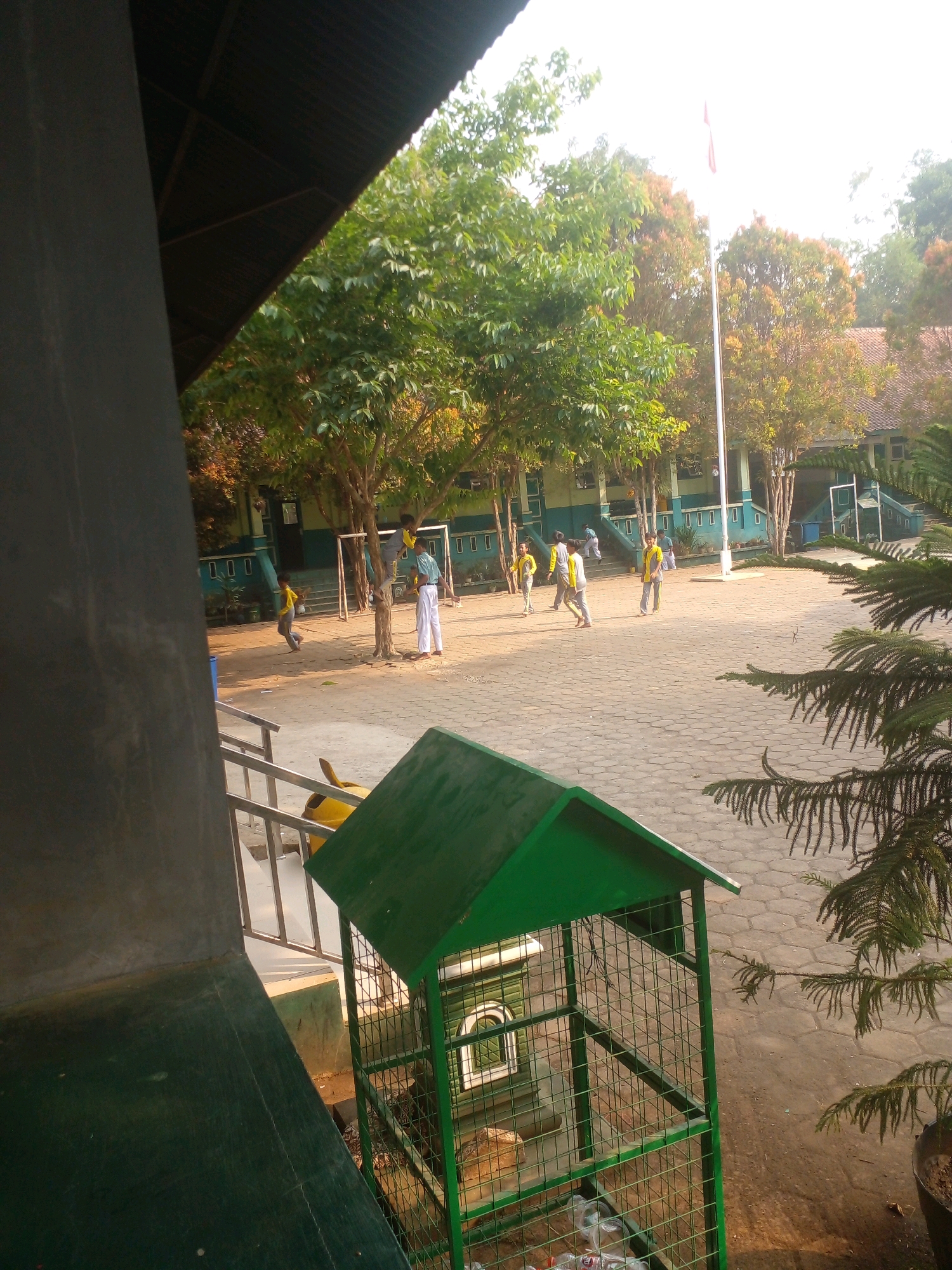 The image depicts a schoolyard scene in Banyuputih, Indonesia, bathed in soft sunlight. In the foreground, a dark, angled structure partially obstructs the view on the left, while a green, open-air trash receptacle with a pitched roof stands prominently on the right. Beyond this, a paved courtyard is populated by several children, appearing to be engaged in recreational activities. Some children are dressed in yellow shirts and dark pants, suggesting a school uniform, and are walking or playing. Two children are near a tree, one standing on what looks like a low wall or structure. In the background, a light-colored building with blue accents forms the school structure, and a tall flagpole with a red and white flag stands near the center. Trees with green and autumn-colored foliage are scattered throughout the scene, contributing to a natural, outdoor environment. The overall impression is a typical school day in a tropical climate.