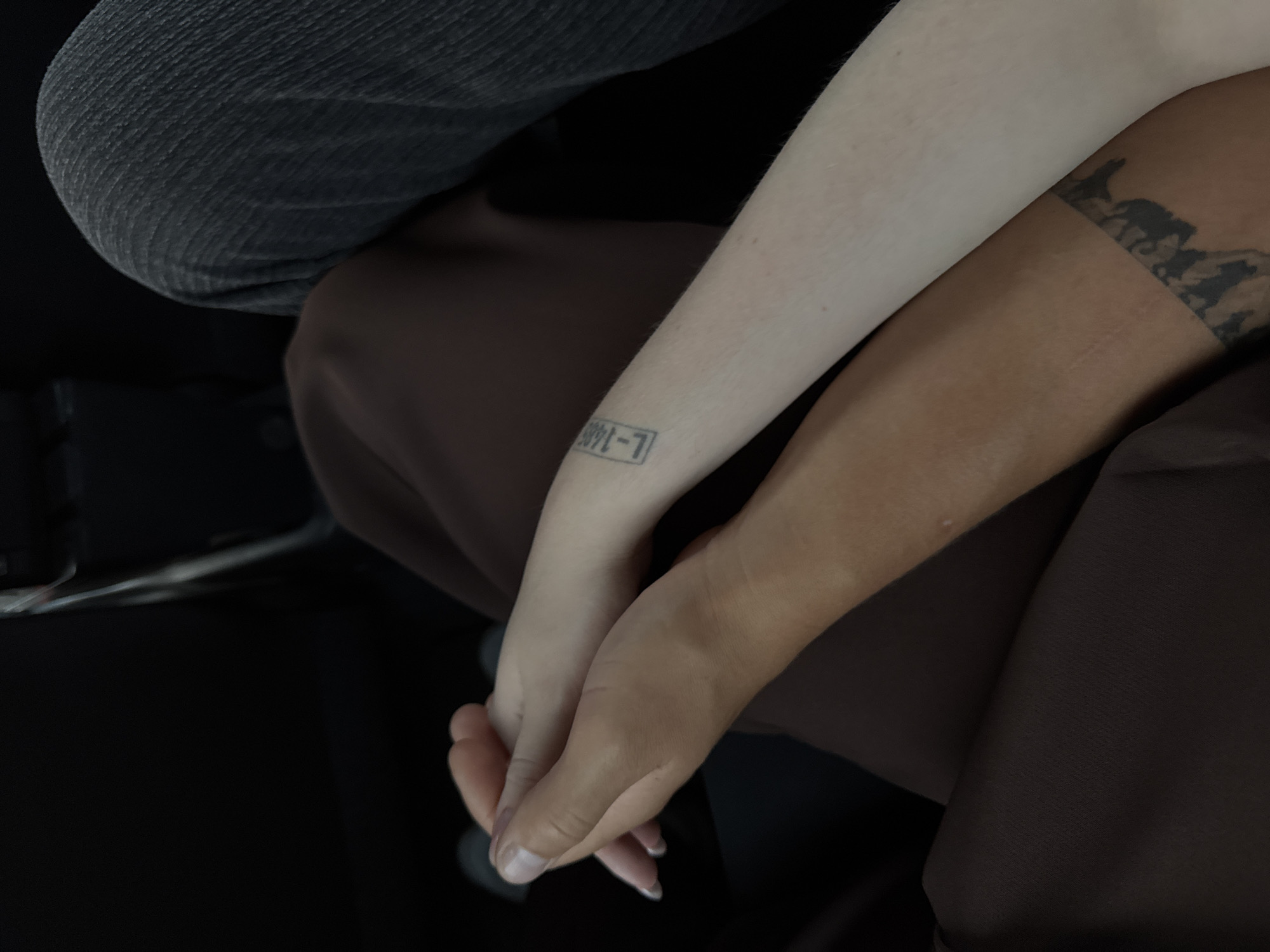 Two individuals are holding hands. One person has fair skin and is wearing a dark brown garment. The other person has tanned skin and is wearing a dark grey denim garment. The fair-skinned individual has a tattoo on their forearm that appears to be a series of characters within a rectangle. The tanned-skinned individual has a tattoo around their forearm that is a repeating pattern of dark shapes. They are seated, suggesting an indoor or vehicle environment.