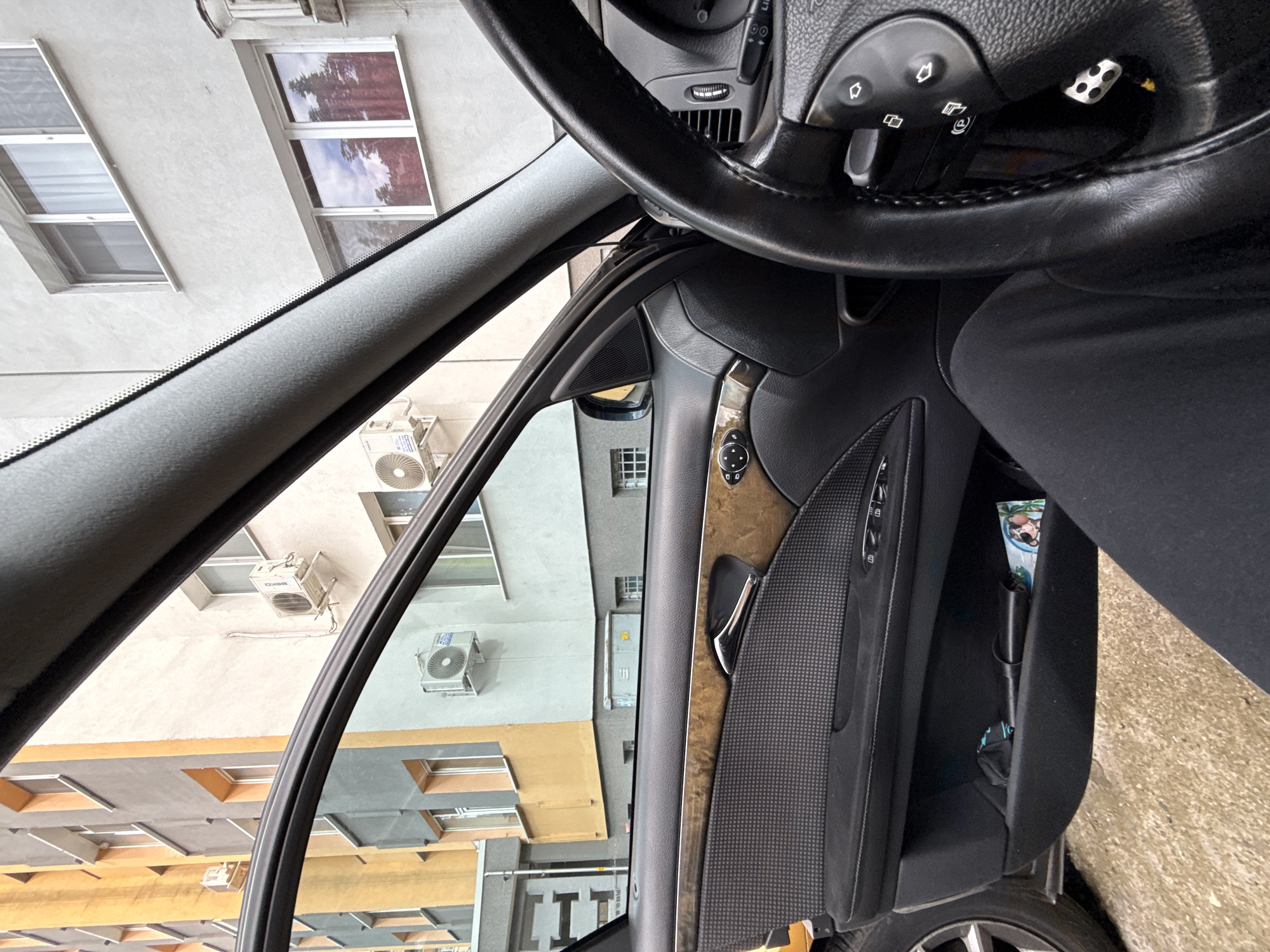 A view from inside a Mercedes-Benz car in Stara Zagora, Bulgaria, focuses on the driver's side door and the exterior scene visible through the window. The black steering wheel, with visible controls, is in the upper right. The door panel is a mix of wood trim, black leather, and textured fabric. Outside, a building with windows and several air conditioning units mounted on its exterior walls can be seen. The buildings suggest an urban environment, indicative of Stara Zagora. There is one individual partially visible in the driver's seat.