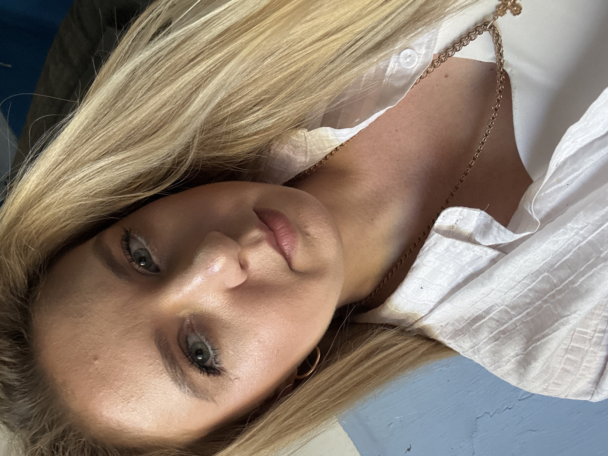 A young woman with blonde hair and green eyes is shown from the chest up, looking at the camera. She is wearing a white collared shirt and a gold chain with a cross pendant. The background is a blue wall. The lighting suggests it is daytime.