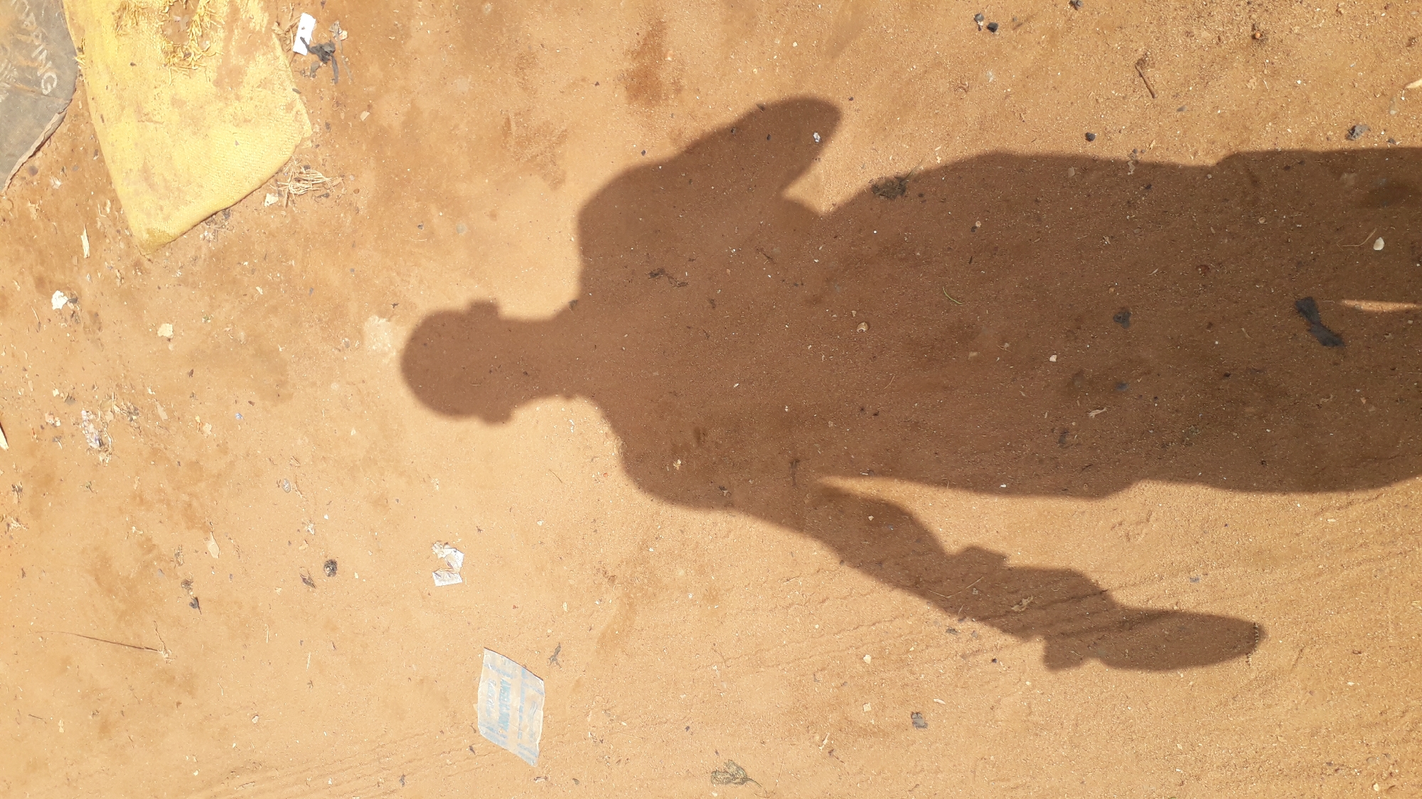 The media file is an image of a shadow of a person cast on sandy ground. The shadow depicts a person standing with one arm outstretched to the side, and the other possibly bent. The ground is a light brown, sandy earth, with some debris scattered on it, including small pieces of paper and dried plant matter. In the upper left corner of the image, a yellow object, possibly a bag or sack, is partially visible. The shadow is long, suggesting that the sun is low in the sky, either early morning or late afternoon. There is no discernible weather condition, but the long shadow implies clear skies. The scene appears to be outdoors, possibly in a dusty or undeveloped area, given the sandy ground and scattered debris. There is no specific event taking place, as the image is a static representation of a shadow. No text is clearly visible in the image.