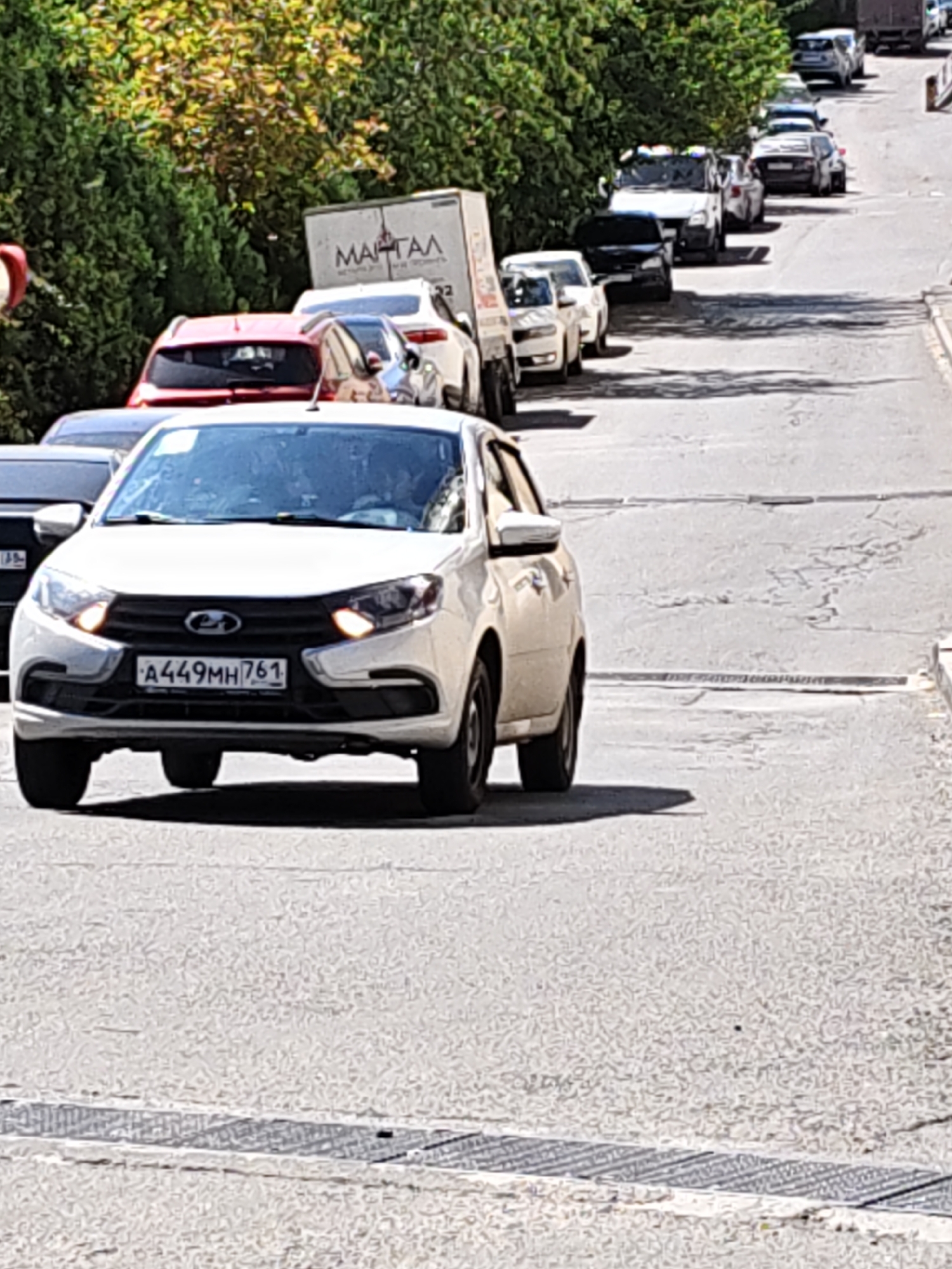 The image shows a street scene in Rostov-on-Don, Russia, on a sunny day. A white Lada Granta sedan with license plate A449MH 761 is in the foreground, facing the camera. The car's headlights are on. Behind it, a line of cars, including a red hatchback, a white van with "МАРТАЛ" written on it, and several other vehicles, are parked or moving along the street. The street is lined with green trees and appears to be a residential or commercial area. The road surface is asphalt, with some cracks and a metal grate visible. The scene suggests a typical urban environment with traffic congestion.