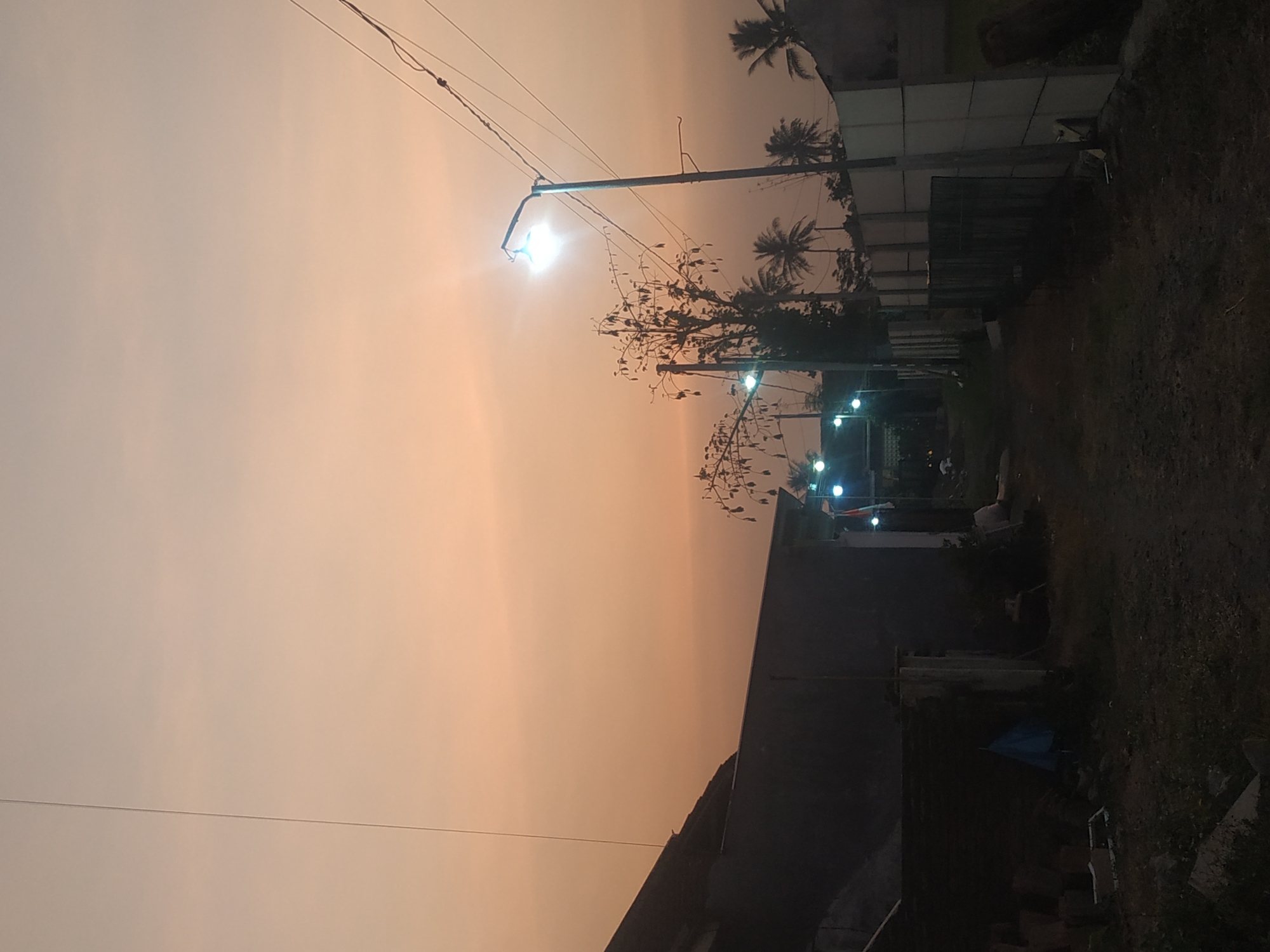 The image depicts a twilight scene in Arjasa, Indonesia, characterized by an overcast, orange sky. A path, bordered by buildings on the left and a fence on the right, leads into the distance. On the left, a dark, silhouetted building with a sloped roof dominates the foreground. To the right of the path, a series of concrete fence panels with vertical supports are visible. A single streetlamp with a bright, illuminated bulb stands prominently on the right side of the path, with power lines stretching across the frame. Several smaller streetlights or decorative lights are also present, illuminating sections of the path and the surrounding area. In the background, silhouetted palm trees and the branches of another tree with hanging seed pods are discernible against the sky. The ground is covered with dirt and some debris. The overall atmosphere suggests a quiet, possibly rural or semi-urban, evening in Arjasa.