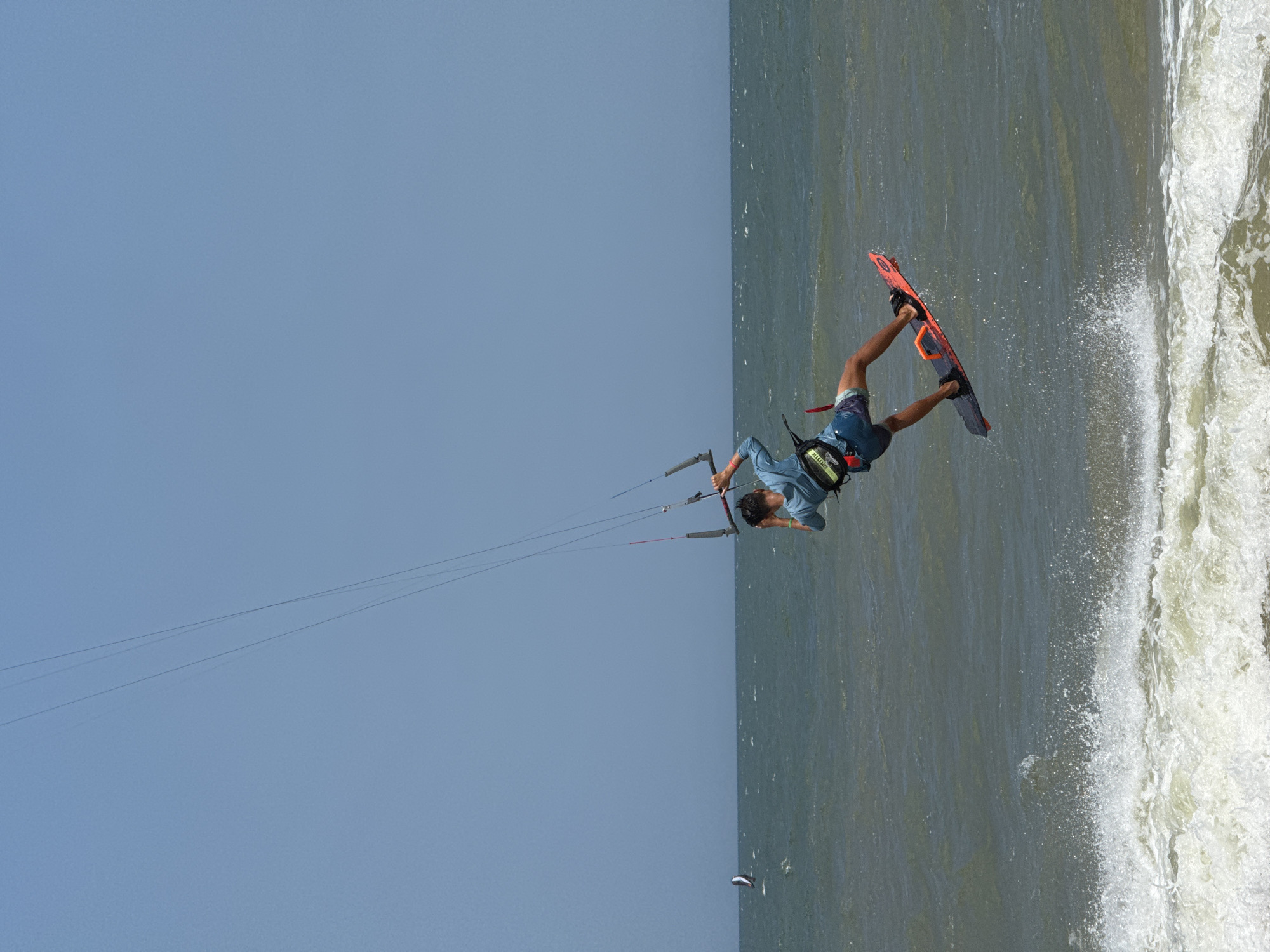 In Mũi Né, Vietnam, on a bright, clear day, a kitesurfer is captured mid-air, performing a jump over the choppy, green ocean waves. The kitesurfer, wearing a blue long-sleeved shirt and patterned shorts, is attached to a kite via control lines and a bar. They are leaning back, balancing on an orange and black kiteboard, with their feet secured in straps. The kite itself is not fully visible, but its lines extend upwards towards the clear blue sky. In the distance, another smaller kiteboarder is visible on the water, also participating in the sport. The scene depicts a moment of athleticism and freedom against the backdrop of the Vietnamese coast.