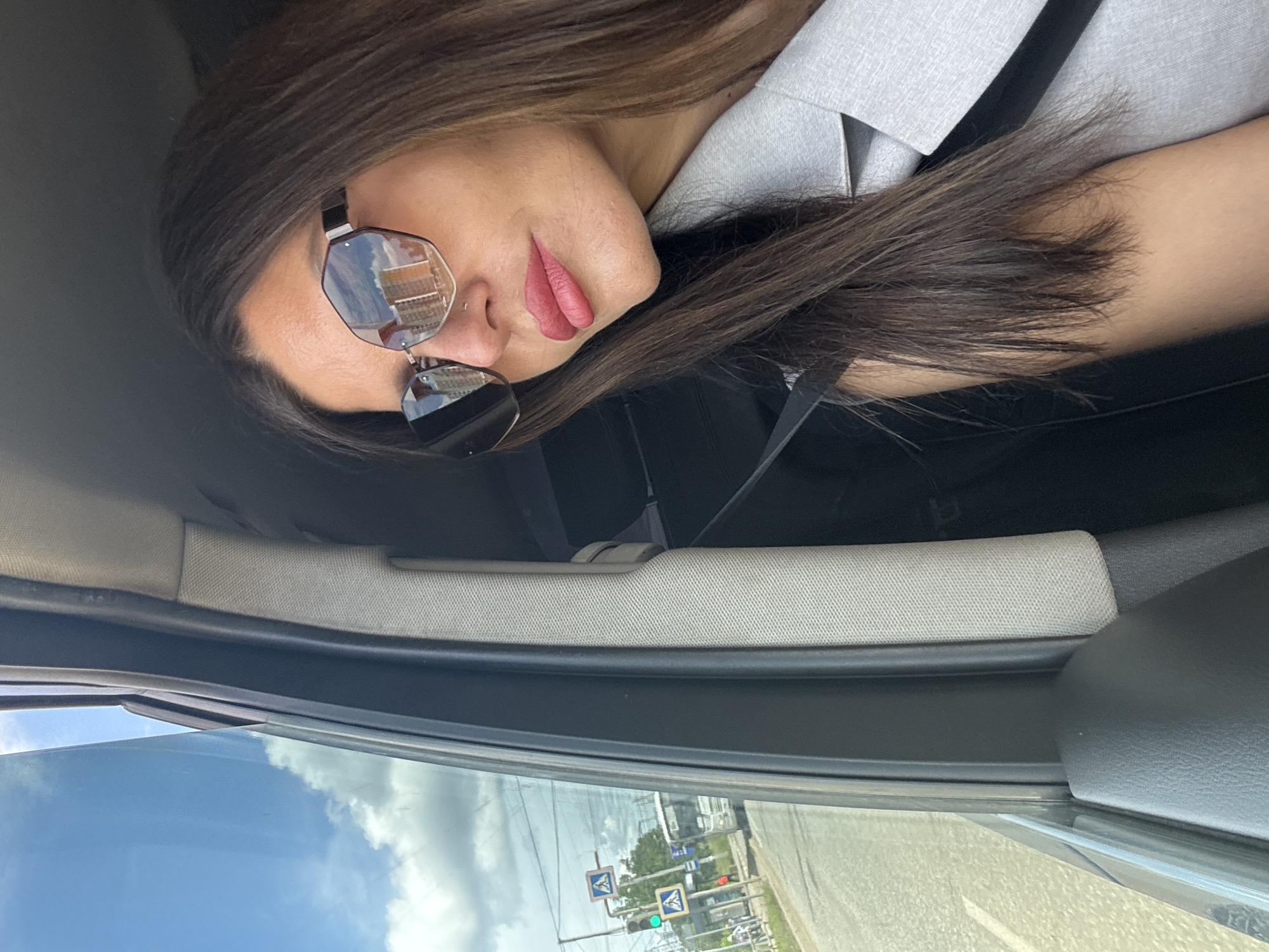 A young woman with long brown hair, wearing sunglasses and a light gray jacket, looks out of a car window. The scene appears to be in Chaikovsky, Russia, as suggested by a street sign reflecting in her sunglasses that bears the Cyrillic alphabet. Outside the car, a street with traffic lights, pedestrian crossing signs, and a bus is visible. The weather is partly cloudy.