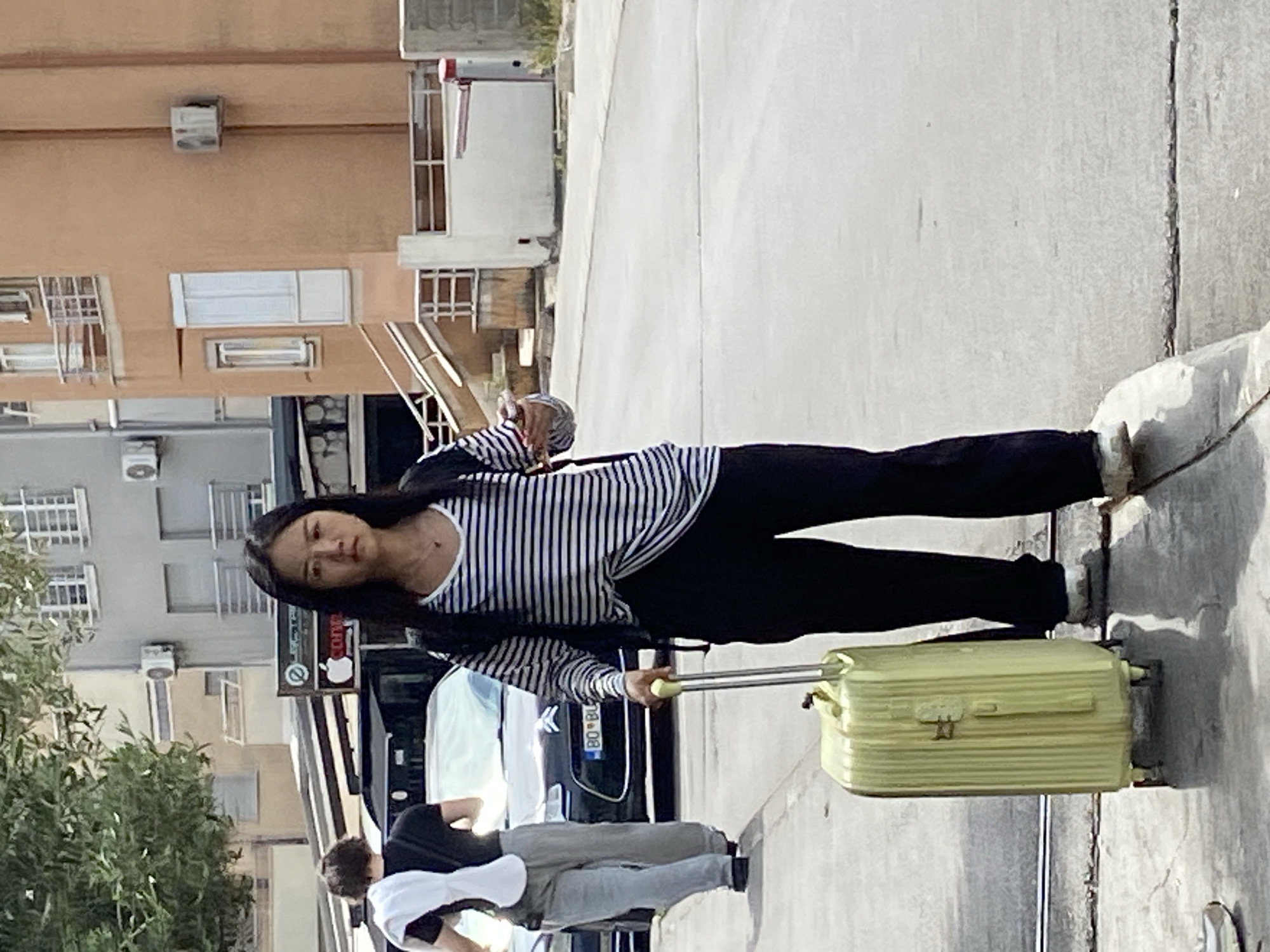 A young woman with long dark hair stands on a sidewalk near the street in what is most likely Budva, Montenegro. She is wearing a black and white striped long-sleeved shirt, black pants, and white sneakers. She is pulling a small, light green suitcase with a silver handle. Behind her, a person wearing a black shirt and light gray pants walks in the same direction. A car is parked nearby. The backdrop includes buildings, trees, and street signs.