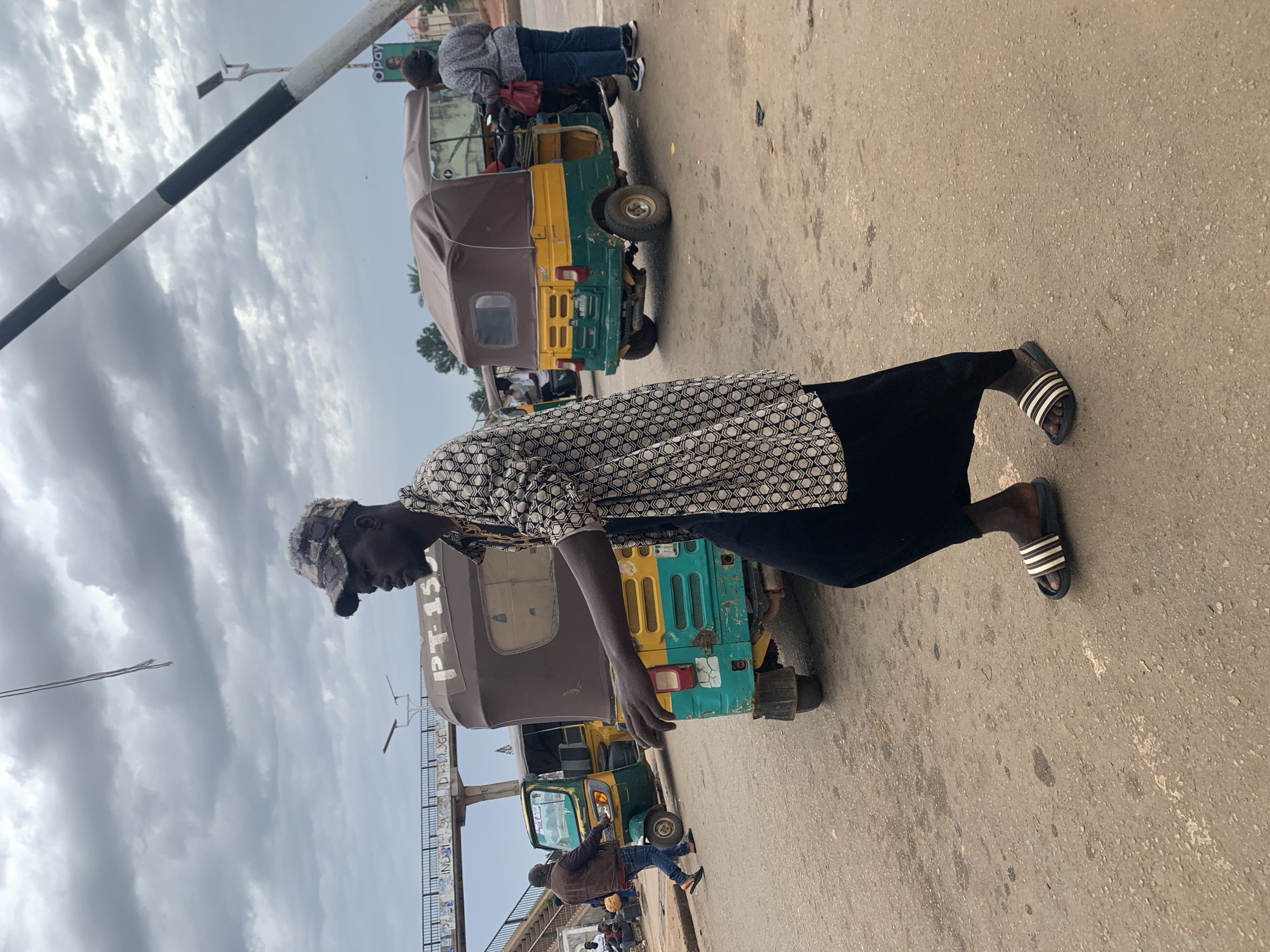 In Gura Top, Nigeria, a man in a camouflage-patterned cap walks on a dusty road. He wears a patterned shirt and a black skirt. He has sandals on his feet. Two auto rickshaws are visible. One is a green and yellow auto rickshaw with the text "PT 15" on its side. Another auto rickshaw is brown and yellow. In the background, a pedestrian bridge is visible and a barrier is mounted on a pole. The sky is cloudy.