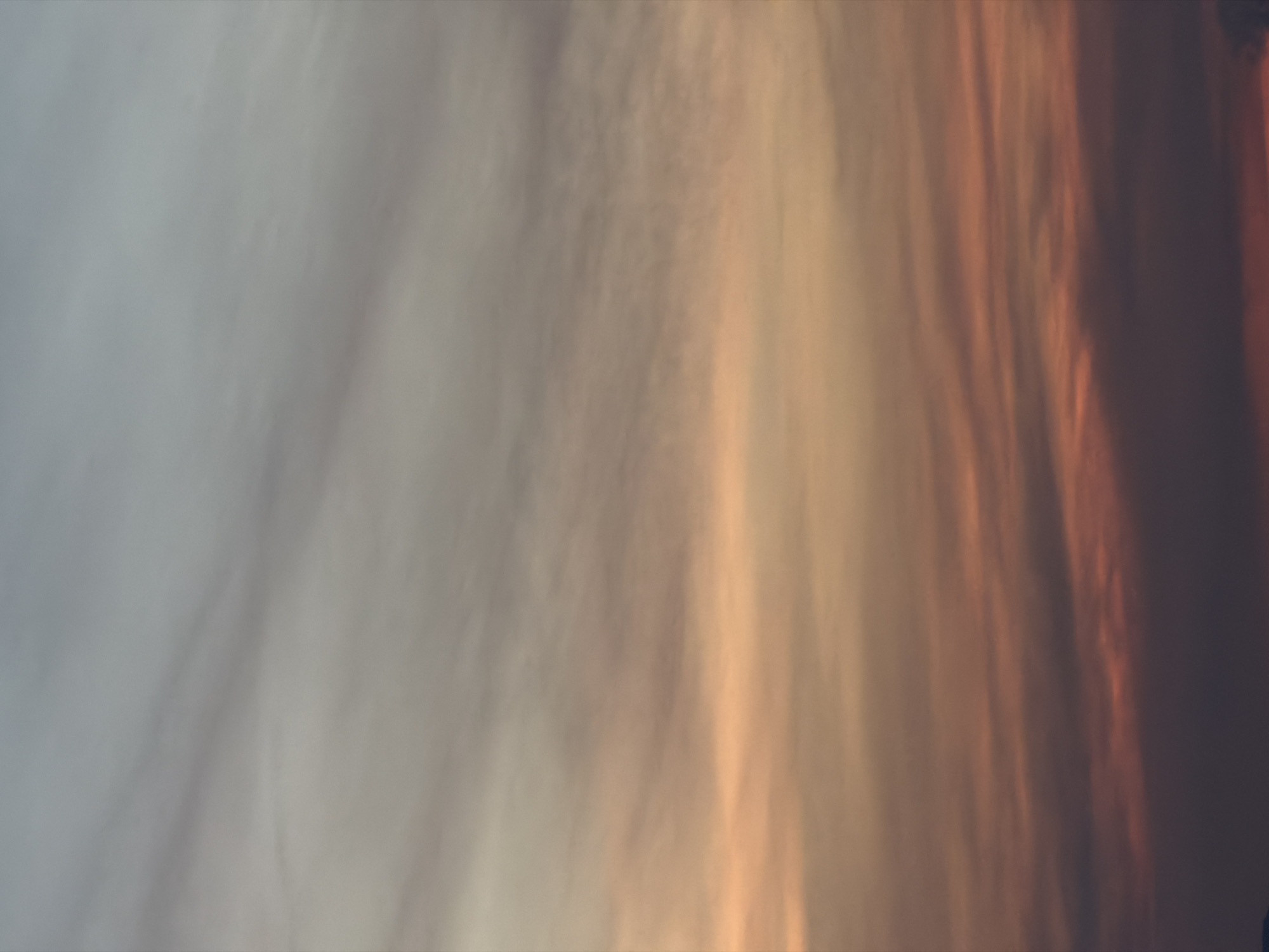 This image shows a sky with wispy clouds at sunset. The colors transition from a light blue-grey at the top to soft orange and pink hues towards the bottom. The clouds are thinly spread and appear to be moving horizontally. The scene is an outdoor environment, likely a wide-open space given the expansive sky. No specific landmarks of Hoi An, Vietnam are visible.