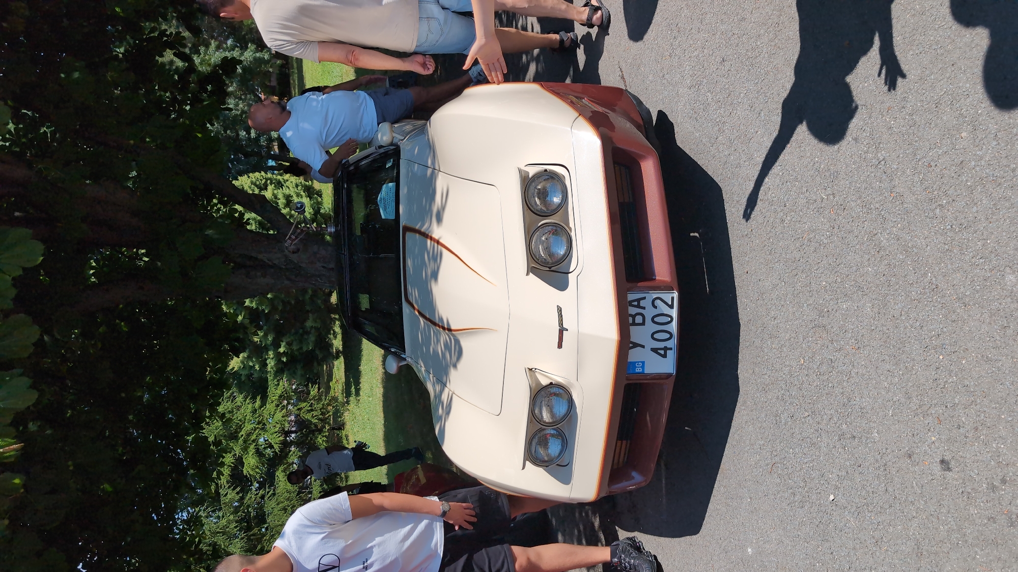 A beige and brown two-door sports car is parked on asphalt, under the shade of a large tree with green leaves. The car has a black convertible top and dual headlights on each side of its front. A license plate with the letters "У ВА" and the numbers "4002" is visible on the front of the car, with a blue and white "BG" sticker in the corner. Several people are standing around the car. One man in a white t-shirt and black shorts stands to the left of the car, while another man in a light blue t-shirt and grey shorts leans on the car to the right. A person wearing a tan t-shirt and denim shorts stands further to the right, with their right hand extended towards the car. The background consists of green grass and more trees. The overall scene suggests an outdoor event or car show. The presence of the "BG" sticker on the license plate indicates the car is registered in Bulgaria. While the exact location within Burgas is not specified, the presence of lush greenery and mature trees points to a park or a well-maintained outdoor public space.