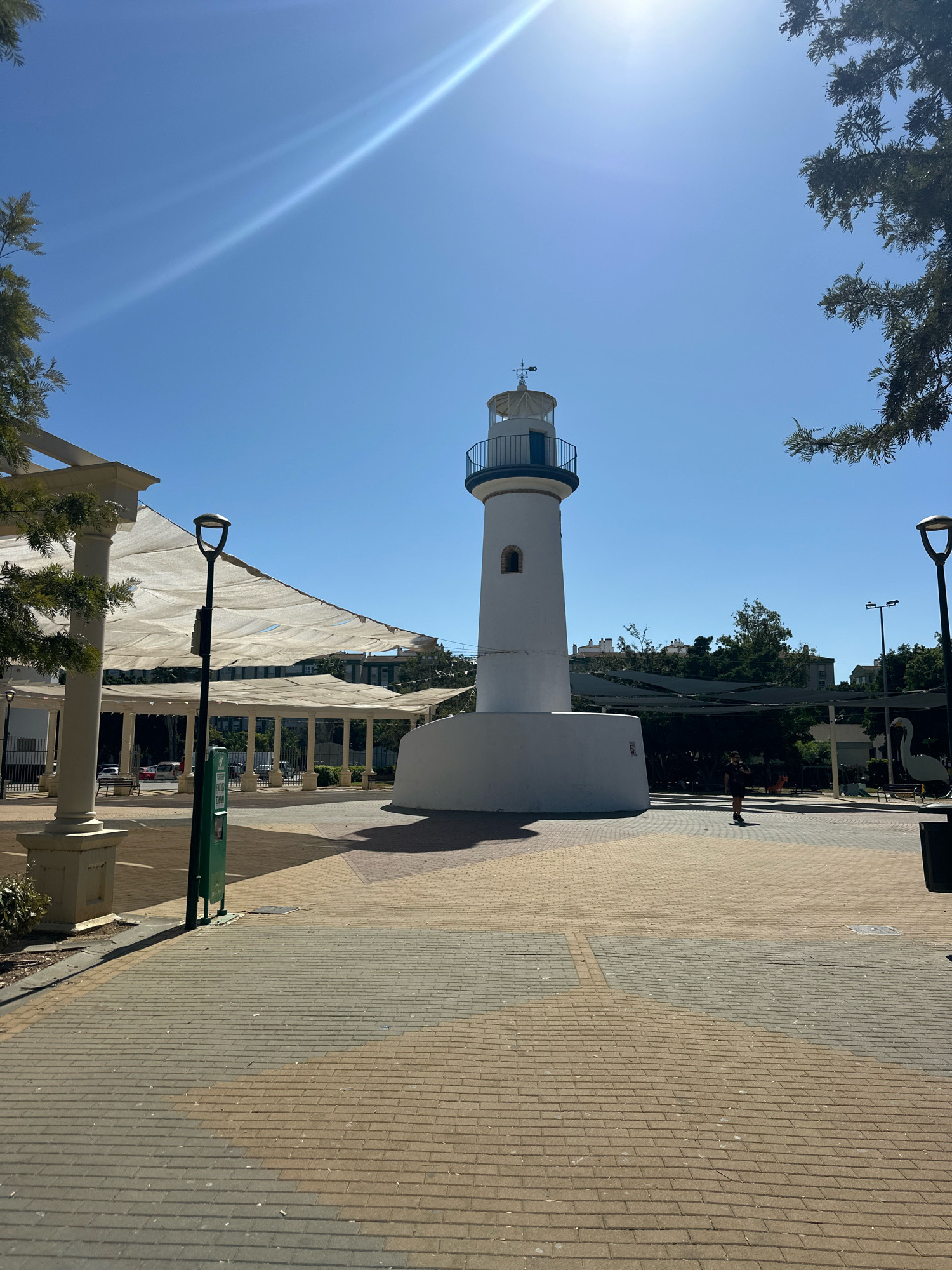 The image shows a white lighthouse in a paved public square in Málaga, Spain. The lighthouse is cylindrical with a white base and a white tower topped with a lantern room. A person is walking in the background. The square is paved with interlocking bricks in shades of brown and grey. There are trees, lampposts, benches, and covered seating areas around the square. The sky is clear and blue with some lens flare from the sun. The lighthouse appears to be a decorative landmark, possibly in a park or promenade. The overall scene suggests a sunny day in an urban outdoor space.