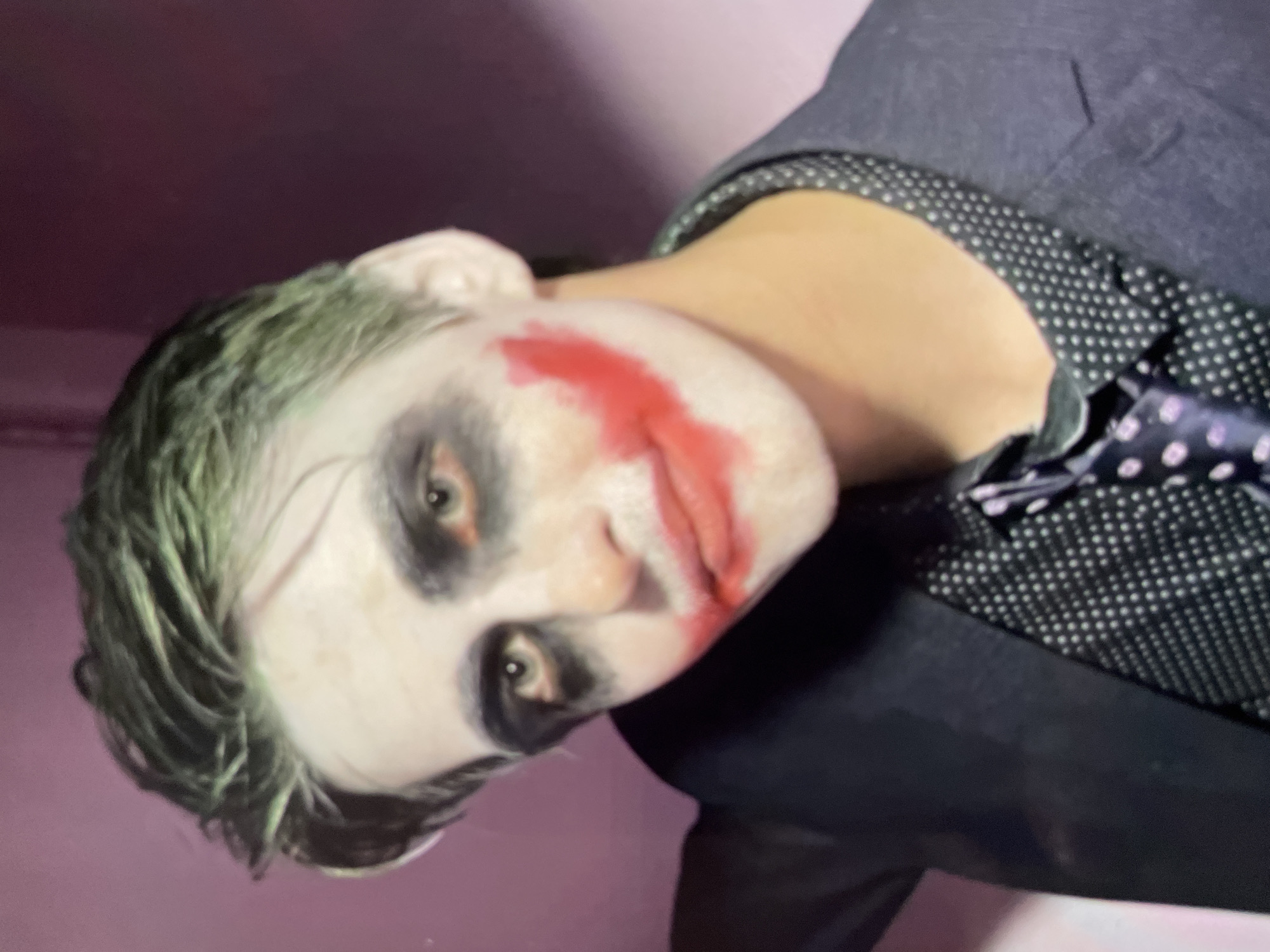 A person with Joker-inspired makeup is seen from a close-up, slightly angled perspective. The makeup includes white face paint, dark circles around the eyes, and red paint smeared to create a sinister smile. The person has greenish hair. They are wearing a dark-colored suit jacket over a polka-dotted shirt and a matching polka-dotted tie. The background is a blurry pink or purple, and there are no other discernible objects or people in the frame. The location in Harmanli, Bulgaria, is unknown.