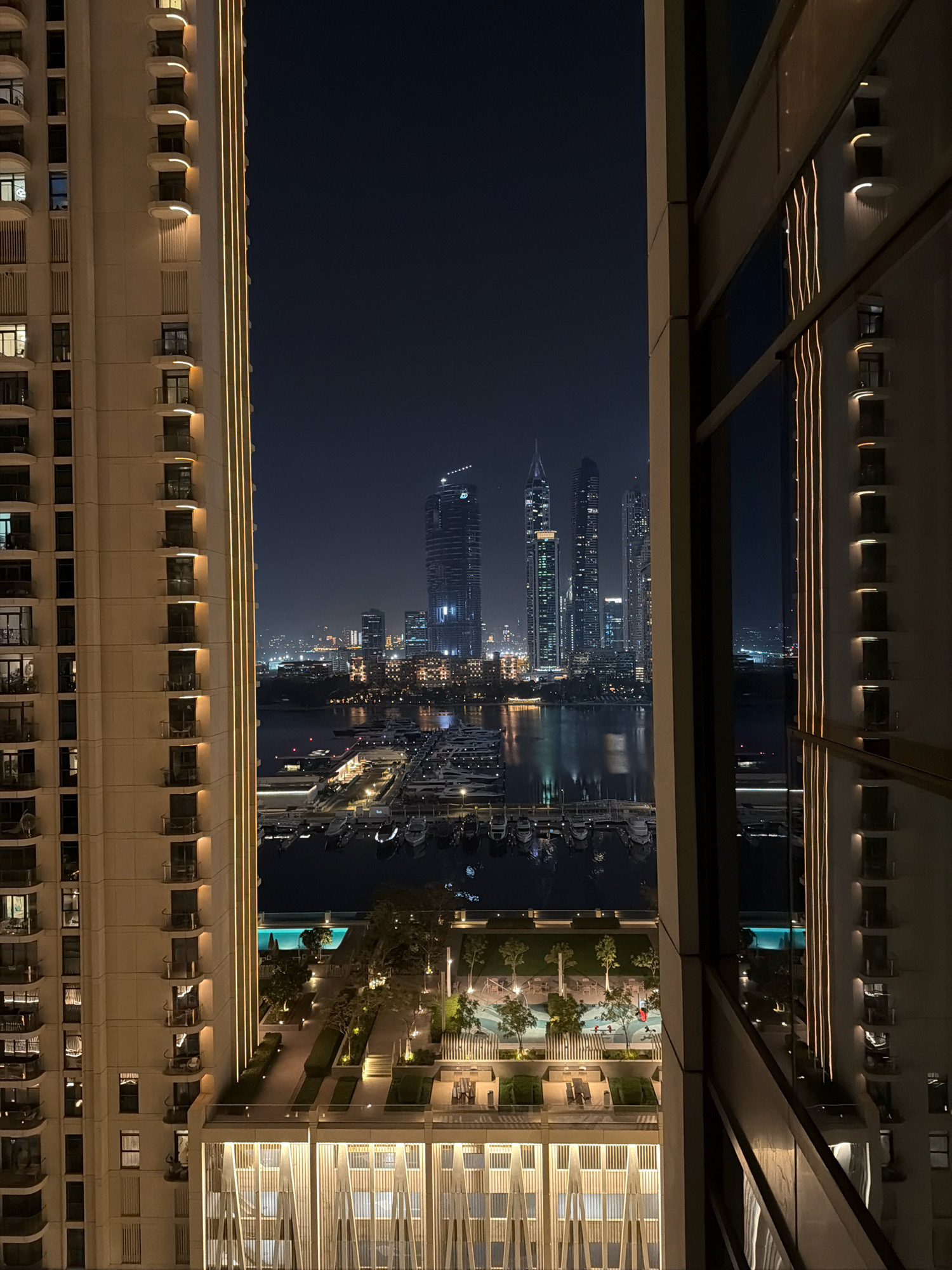 The image captures a nighttime view of the Dubai Marina, United Arab Emirates. On the left, a modern residential building with illuminated balconies and vertical lighting strips stands tall. To the right, another building's facade is visible, with its windows reflecting the exterior lights and the city skyline. In the center, the Dubai Marina is filled with numerous yachts and boats docked at the marina. Beyond the marina, the Dubai skyline is dominated by various contemporary skyscrapers, their lights twinkling in the dark sky. A tranquil body of water reflects the city lights and the surrounding structures. Further in the foreground, there's an outdoor recreational area with a swimming pool, lush greenery, and seating areas, all illuminated by ambient lighting. The overall scene depicts a vibrant urban waterfront at night, characterized by modern architecture and bustling maritime activity.
